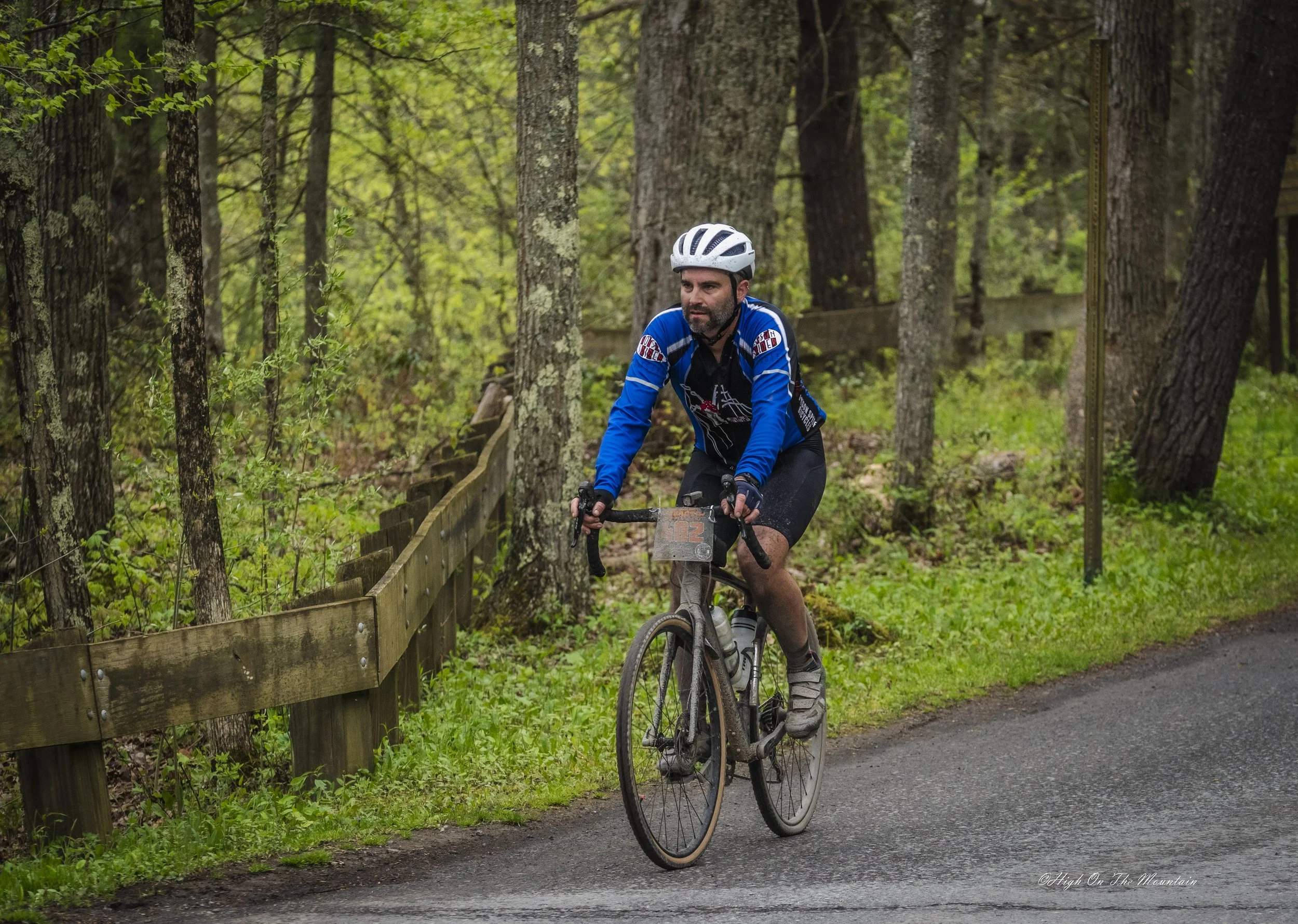 A man riding a mountain bike on a trail in a dense green forest, wearing a blue cycling jacket, black shorts, gloves, and a white helmet.
