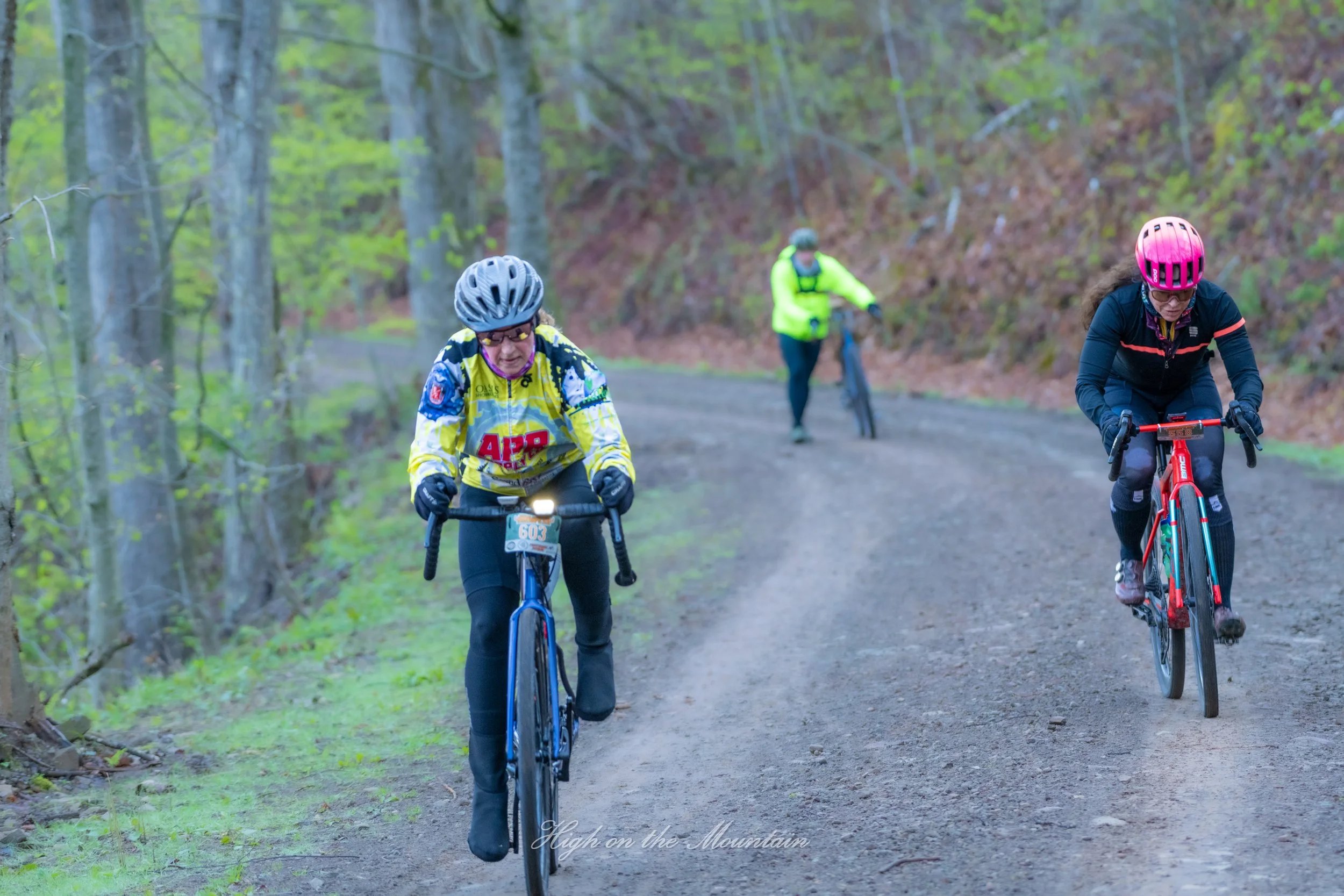 Three women mountain biking on a dirt trail through a forest with green foliage, wearing helmets and riding gear.