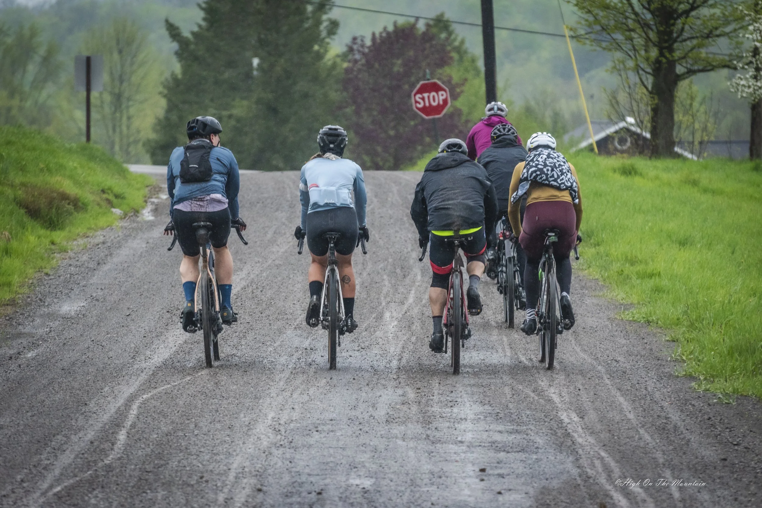 A group of six cyclists riding on a gravel road surrounded by green grass and trees, with a stop sign and power lines in the background.