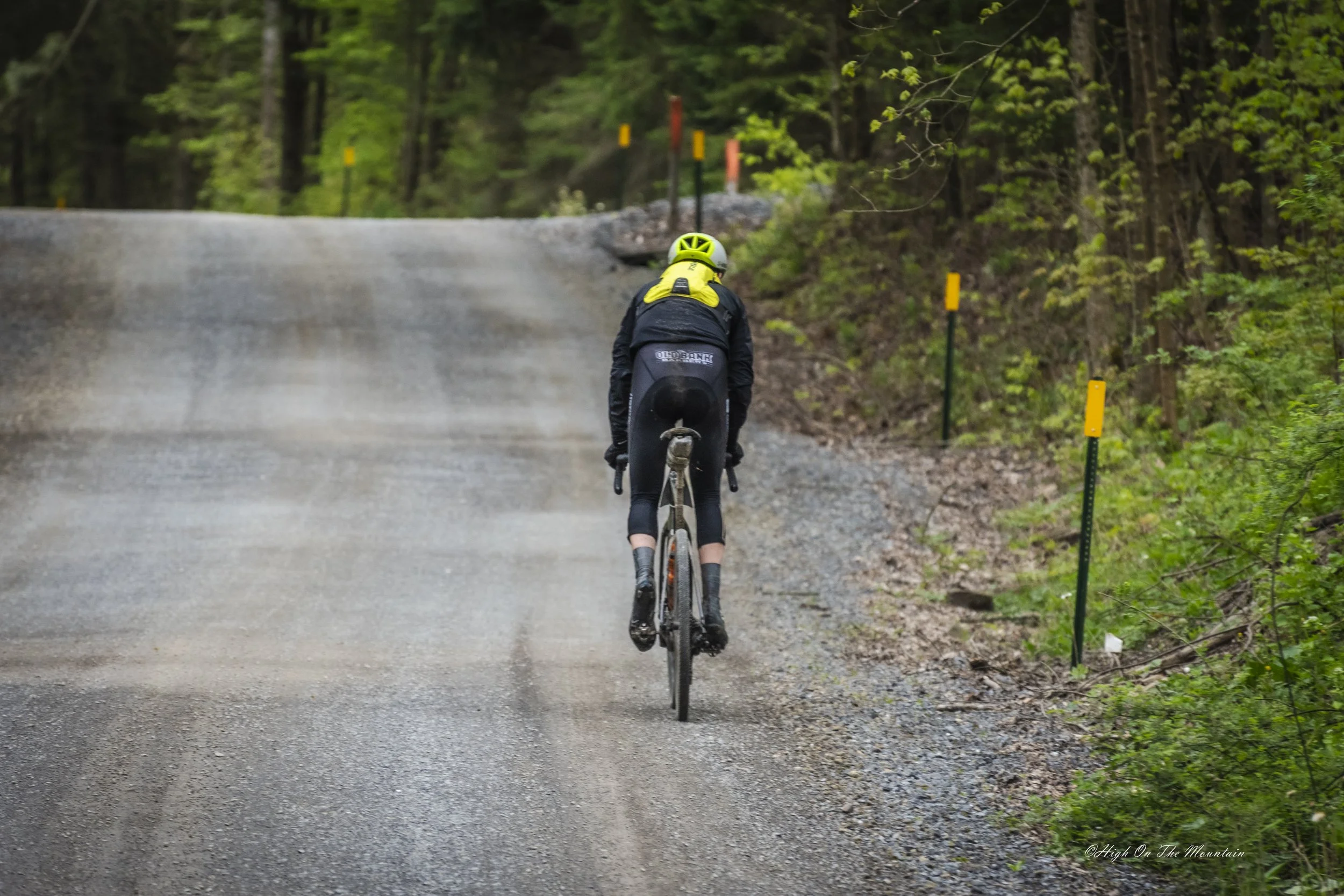 A person riding a bicycle on a gravel forest trail surrounded by trees and green foliage, with yellow and orange trail markers along the side.