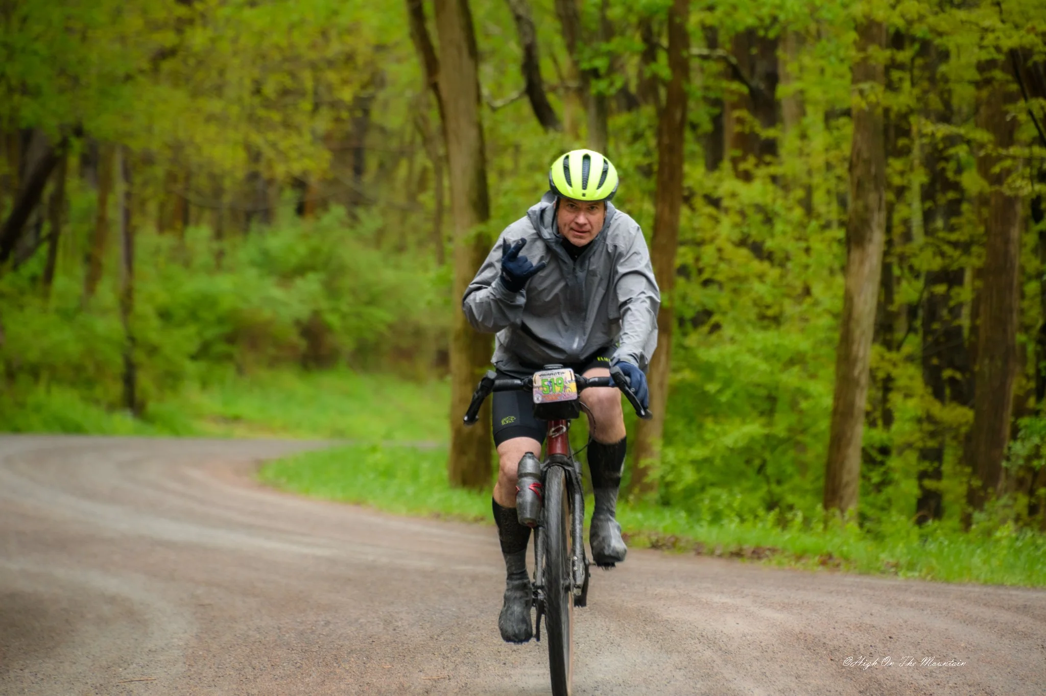 A man wearing a gray jacket, yellow helmet, and gloves riding a mountain bike on a forest trail surrounded by green trees.