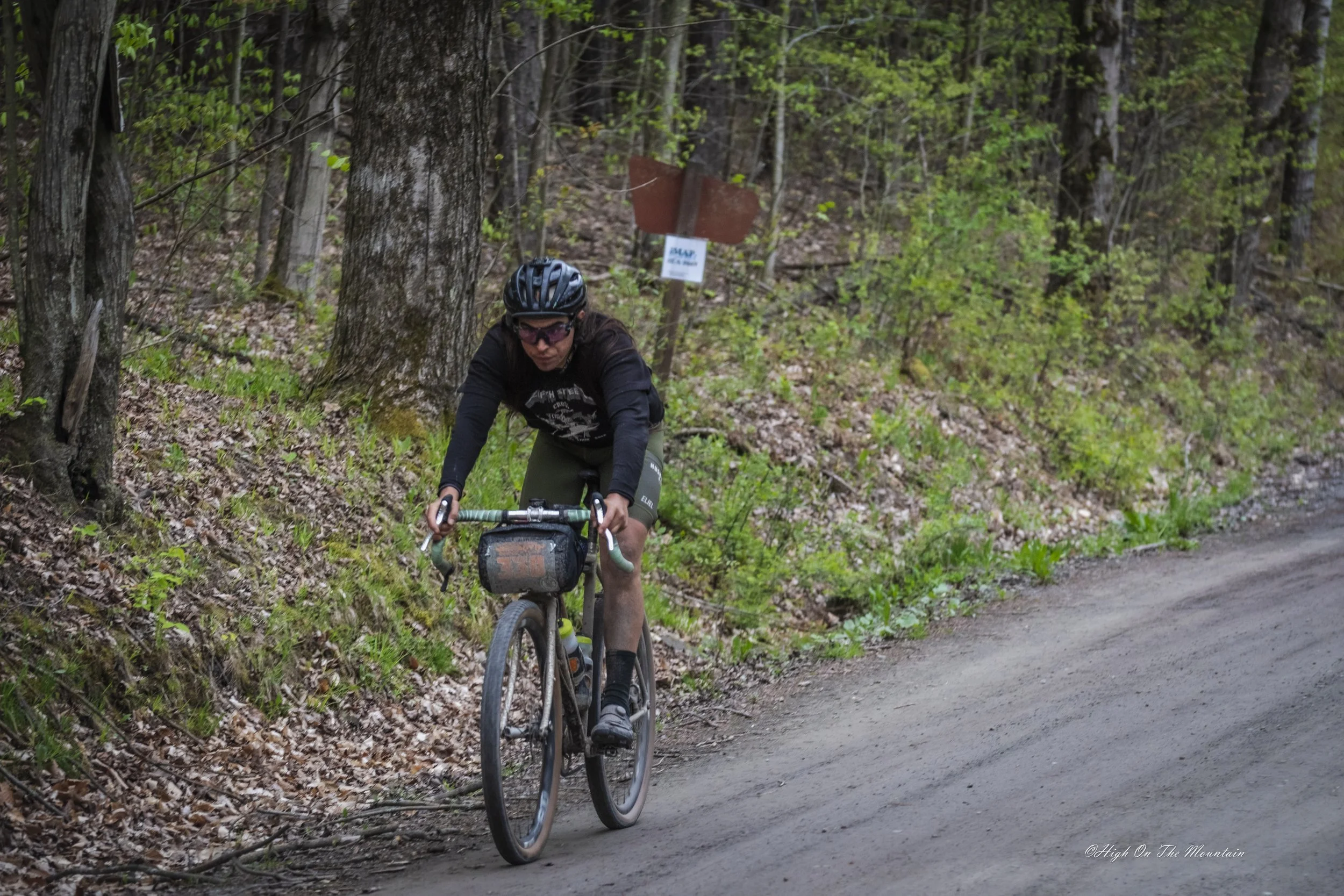 A person wearing a black helmet, sunglasses, a black long-sleeve shirt, and green shorts riding a gravel bike on a dirt trail in a forest with green foliage.