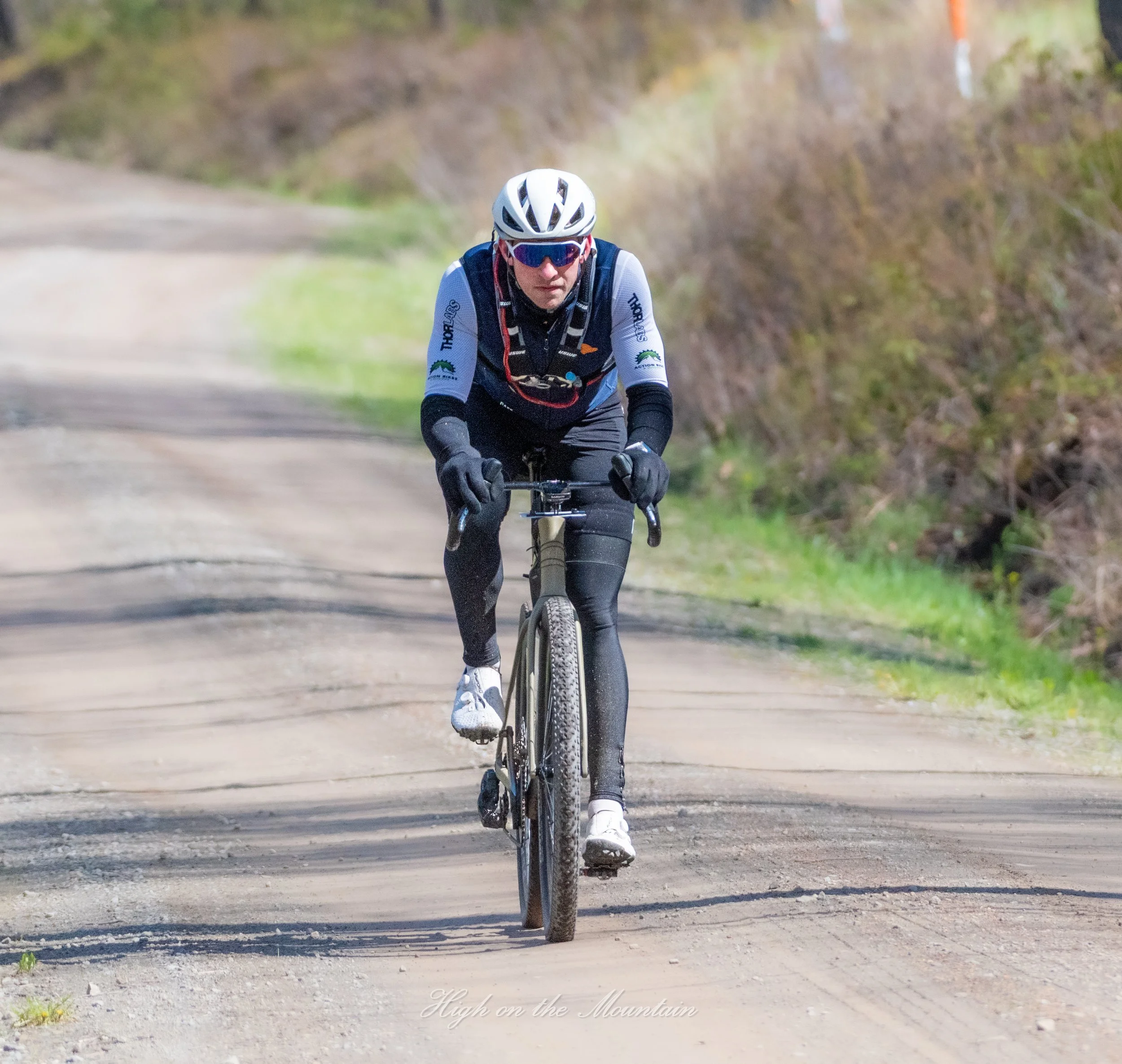 A man wearing cycling gear, a white helmet, and sunglasses riding a mountain bike on a dirt trail through nature.