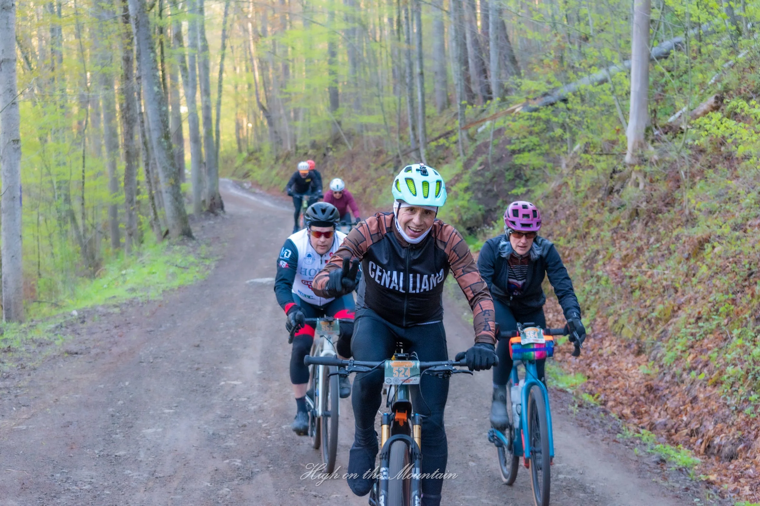 A group of mountain bikers riding on a dirt trail through a lush green forest, wearing helmets and protective gear.