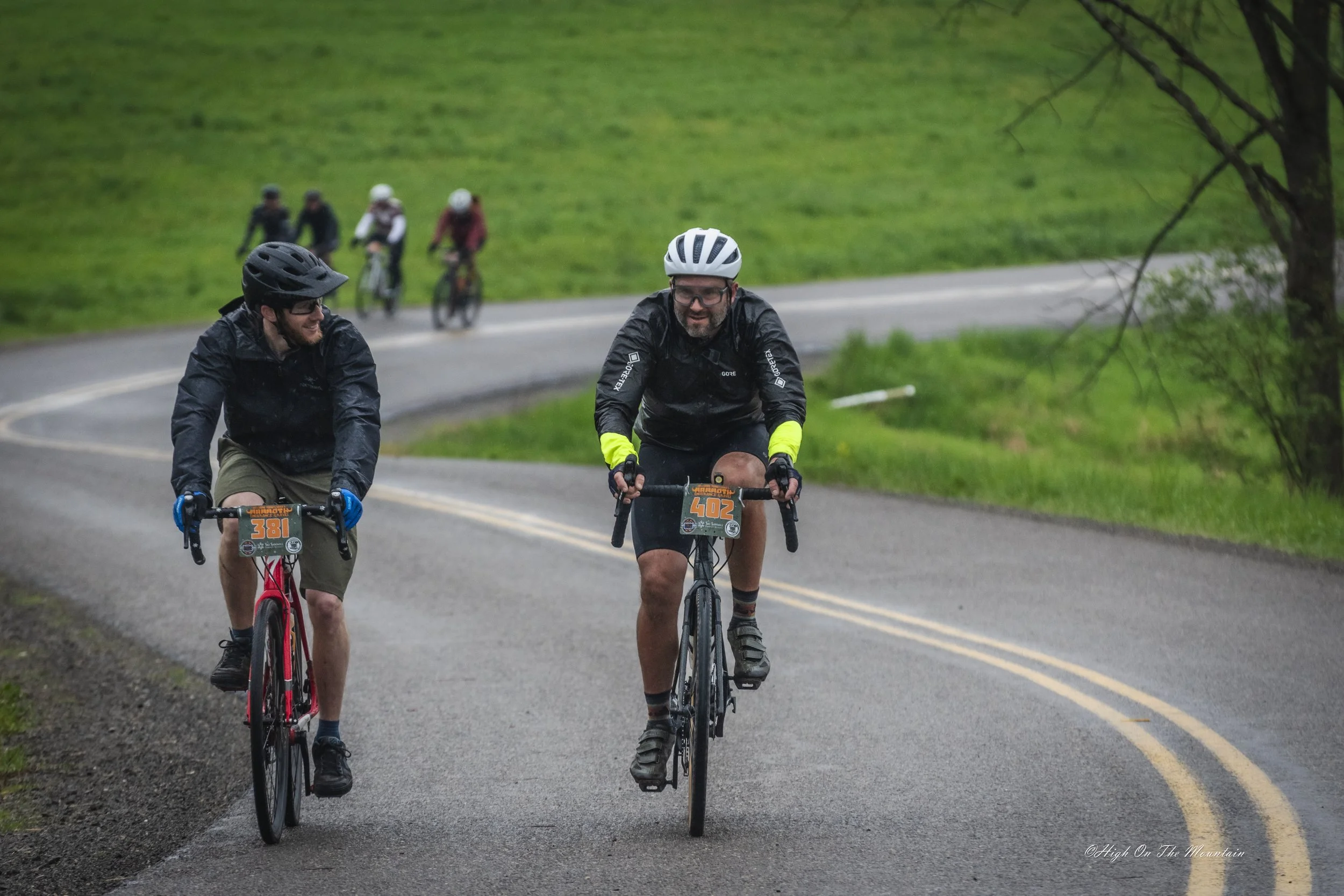 Two men riding bikes on an outdoor road with a green hill and four additional cyclists visible in the background, all wearing helmets and raincoats.