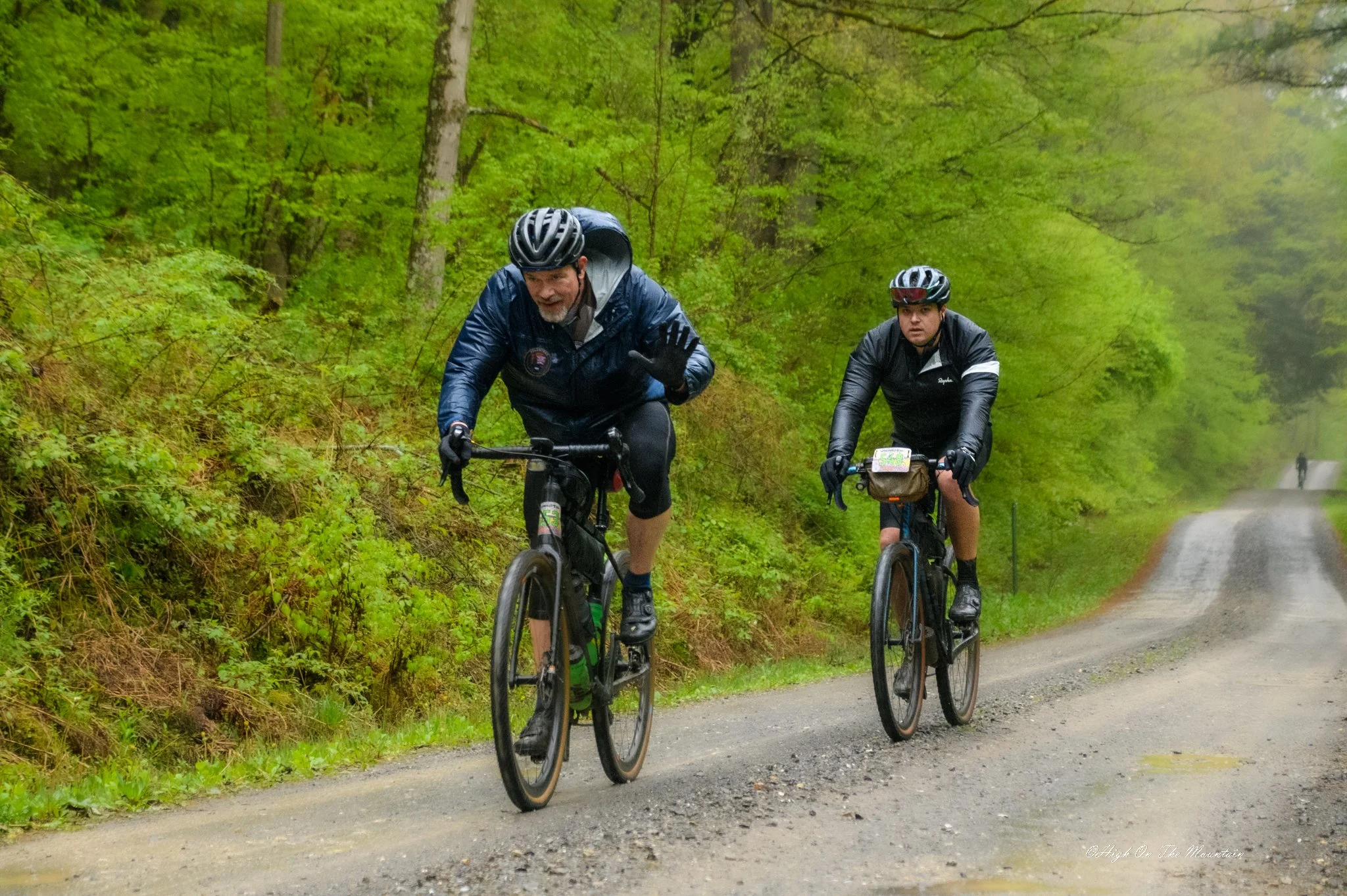 Two men riding mountain bikes on a trail through a lush green forest with misty weather.