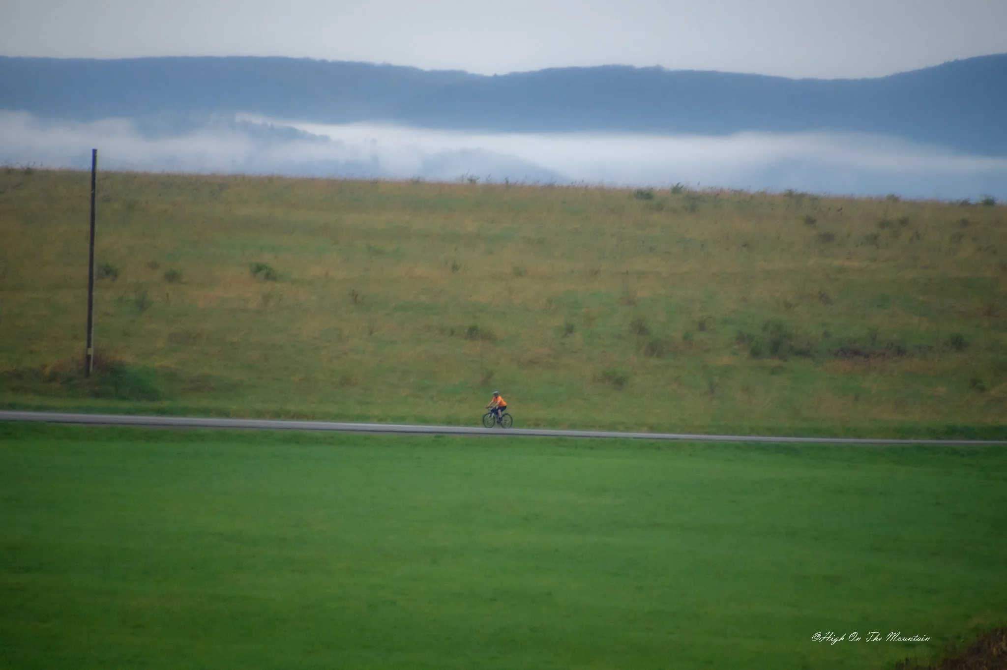 A lone cyclist wearing an orange shirt riding on a flat rural road with green grass in the foreground, a grassy hill with a blue pole, and fog-covered mountains in the background.