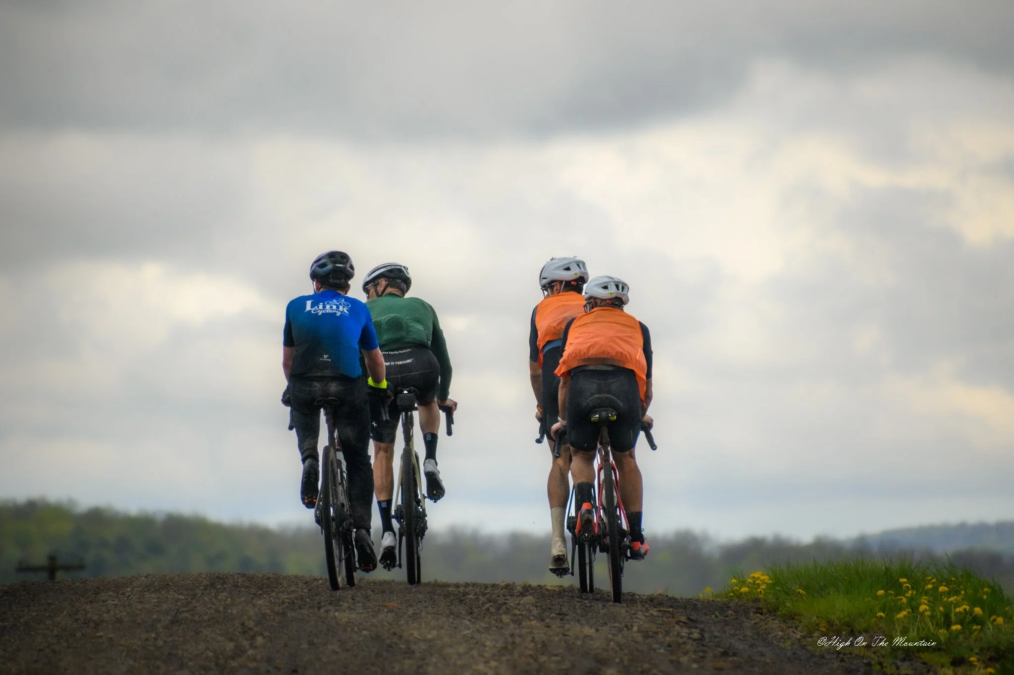 Four cyclists riding on a dirt path in an open landscape under cloudy sky.
