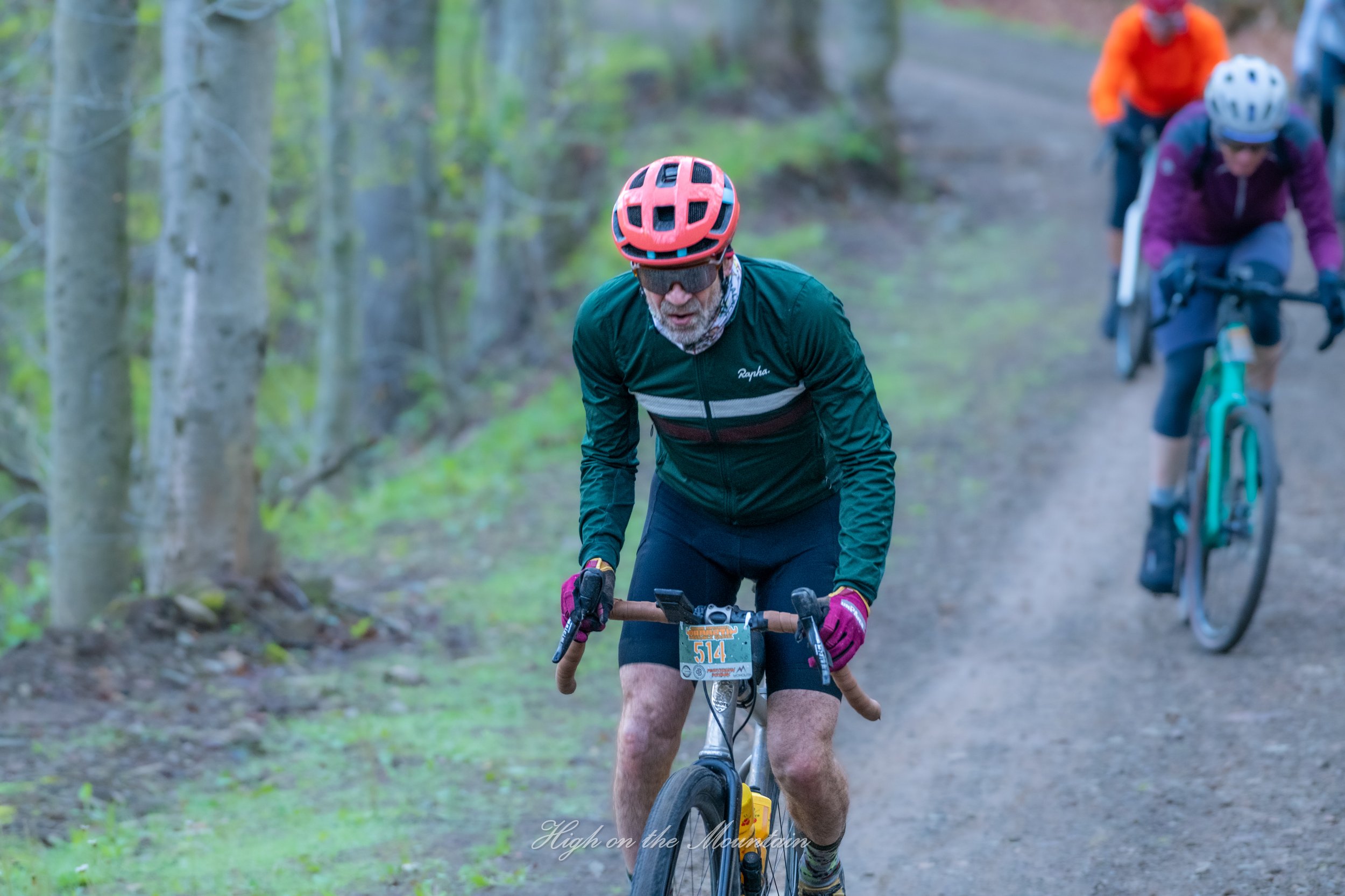 A group of mountain bikers riding along a forest trail, with one man in the foreground wearing a red helmet and sunglasses, leading the pack on a bike. The scene is set in a lush, green forest with tall trees on either side of the trail.