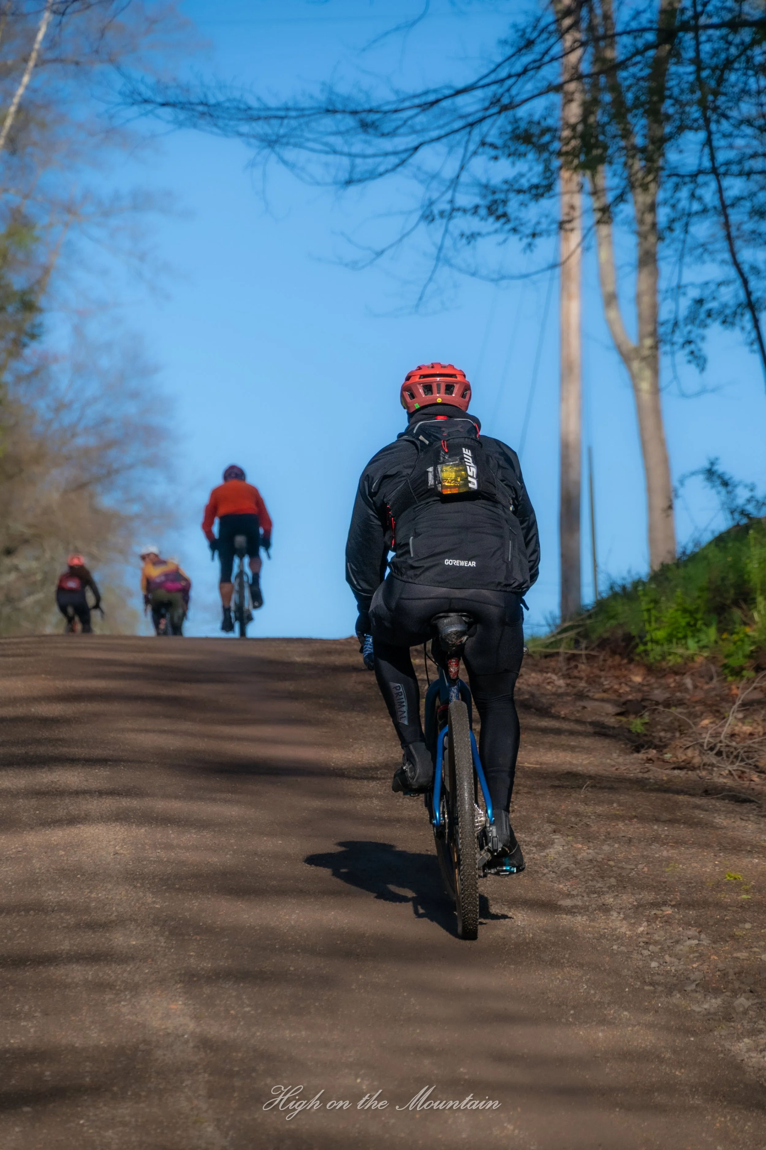A group of cyclists riding on a dirt trail through a forested area during dusk or dawn. The image shows the back of a cyclist in front, wearing a red helmet and black gear, with other cyclists ahead. The sky is a deep blue, and the scene is surrounde