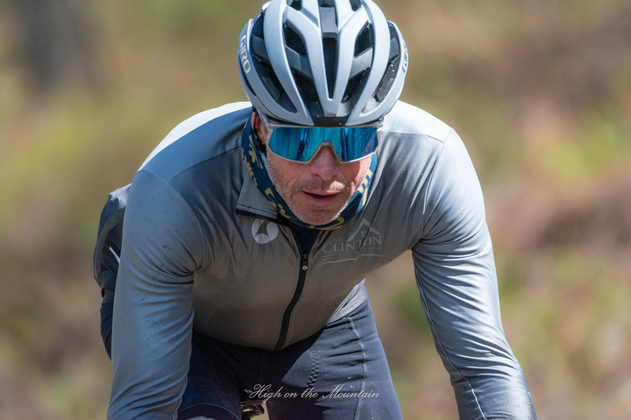 A man riding a mountain bike outdoors, wearing a bike helmet and sunglasses, with a blurred natural background.