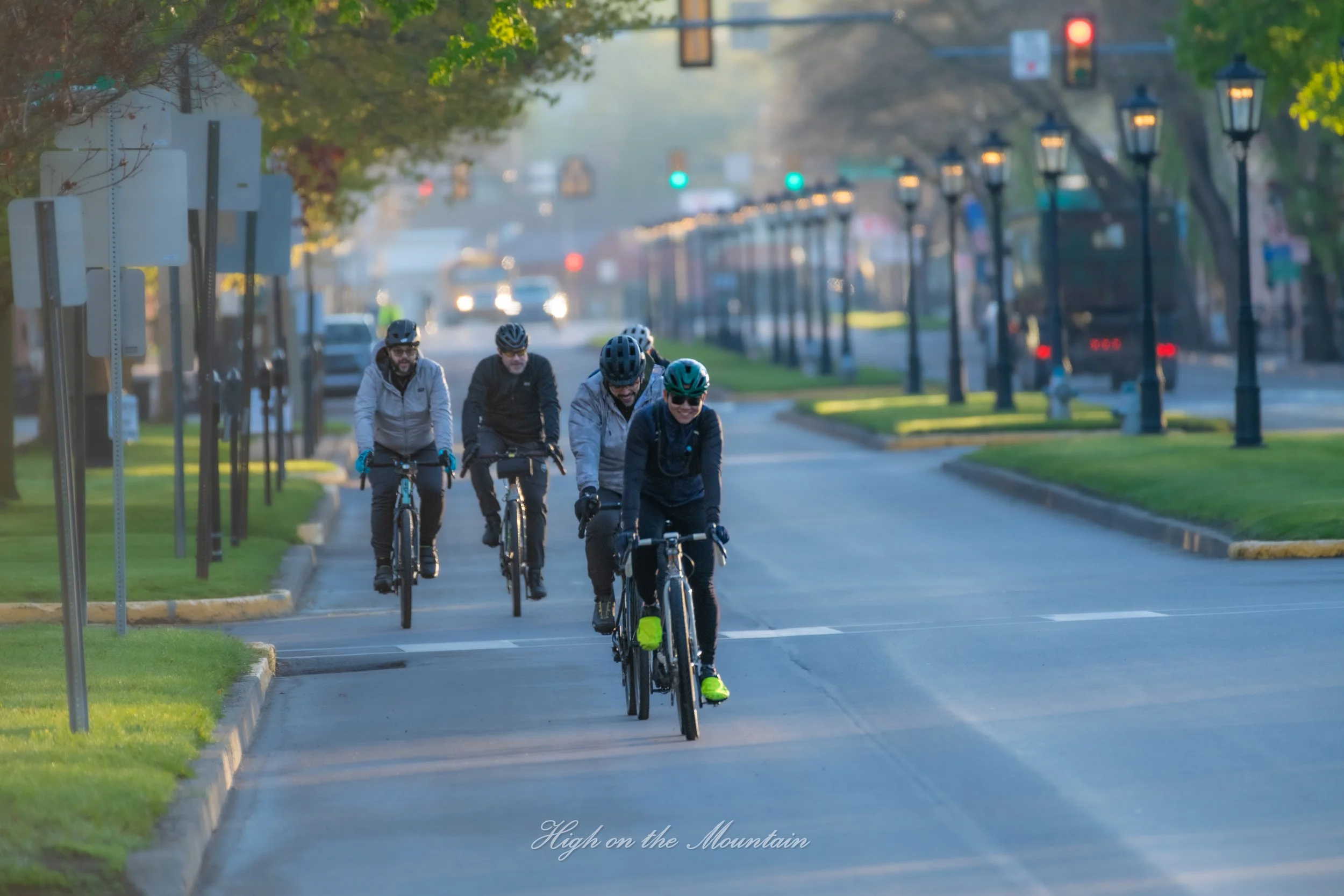 Four men riding bicycles on a city street during early morning or late afternoon, wearing helmets and outdoor gear, with green traffic lights ahead, trees lining the sidewalk, and streetlights along the road.