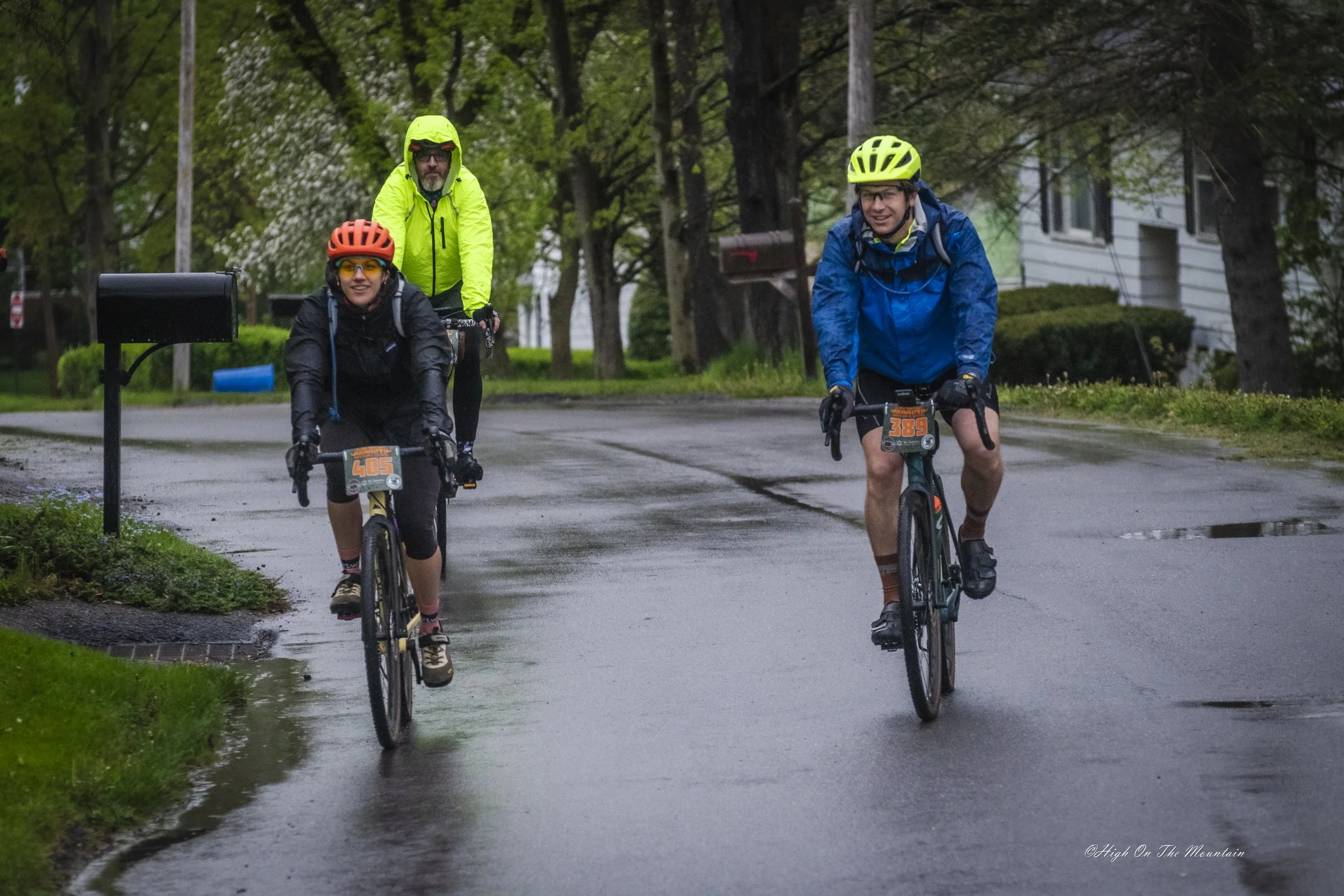 Three cyclists riding on a wet suburban street surrounded by greenery, wearing rain gear and helmets, with a mailbox and residential houses in the background.