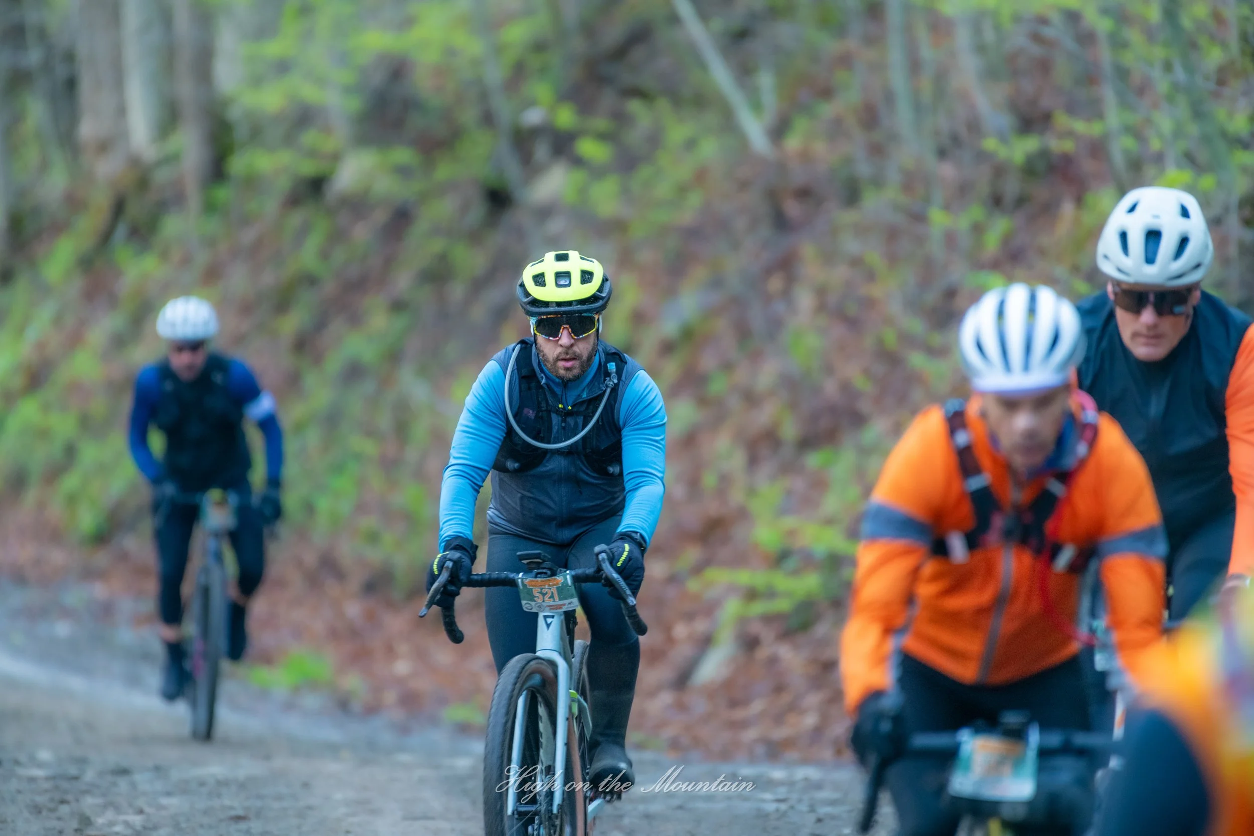 A group of four mountain bikers riding on a trail in a forest with green foliage, wearing helmets and sports gear.