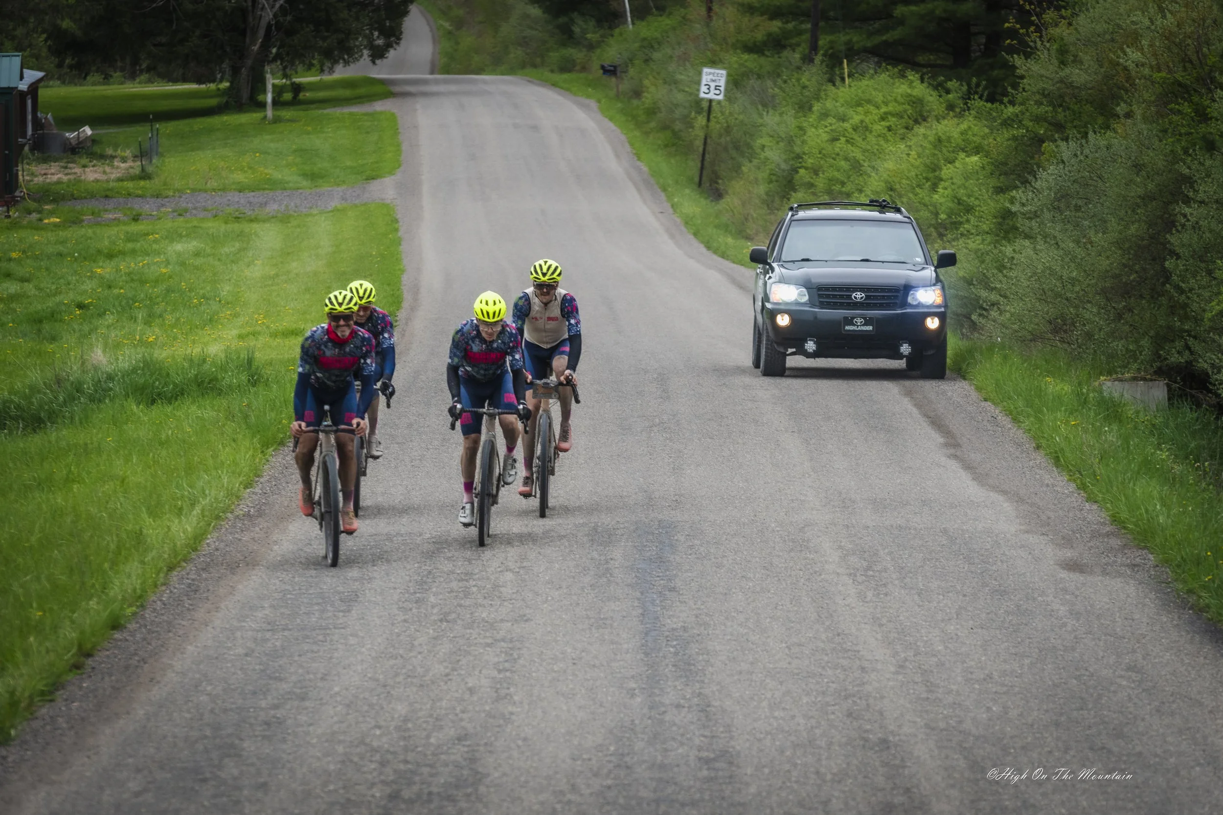 Four cyclists wearing yellow helmets ride on a rural gravel road, with a black SUV parked on the right side and green trees and grass surrounding the area.