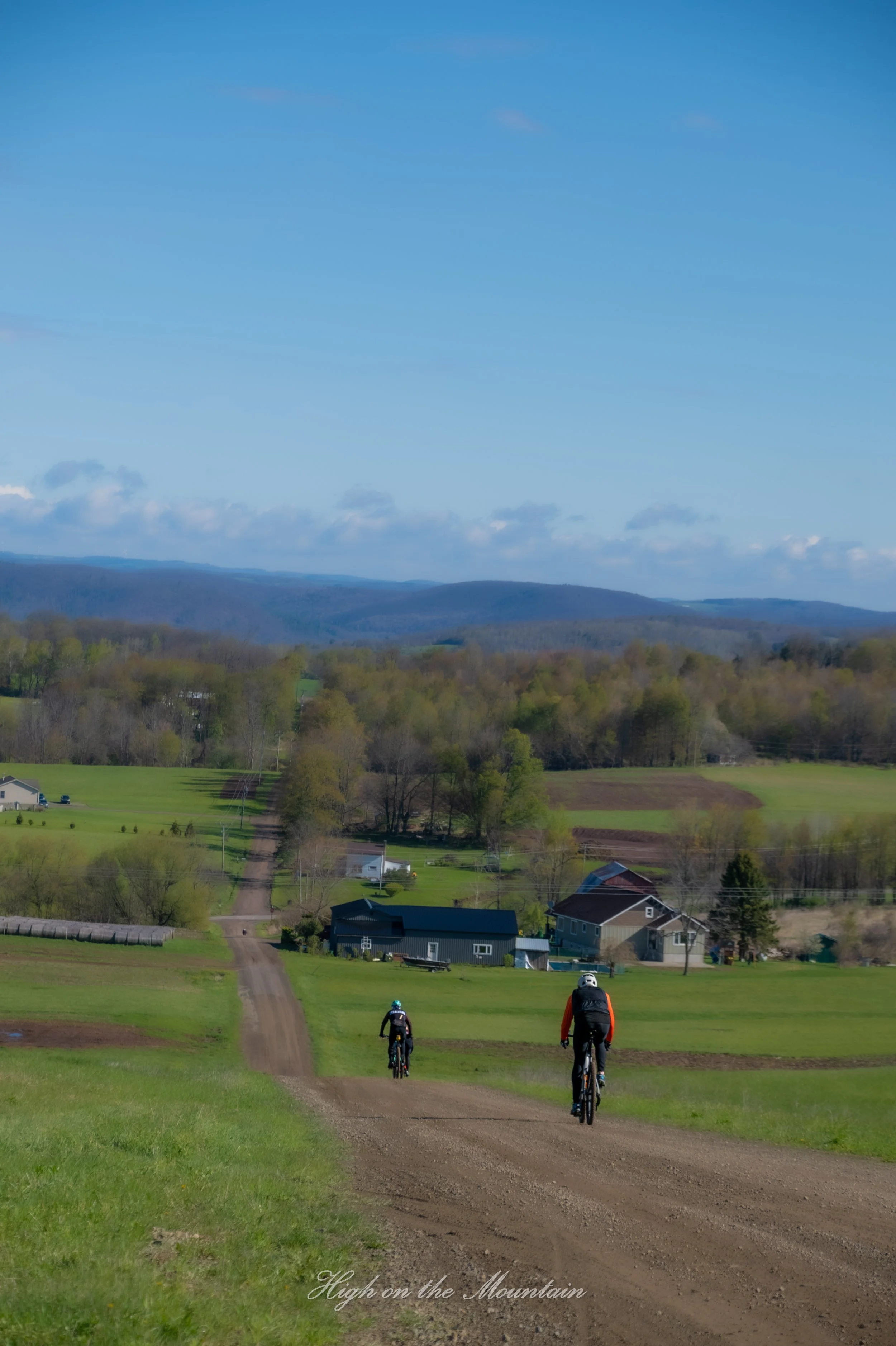 Three mountain bikers riding downhill on a dirt trail through lush green fields toward houses, with hills and a blue sky with clouds in the background.