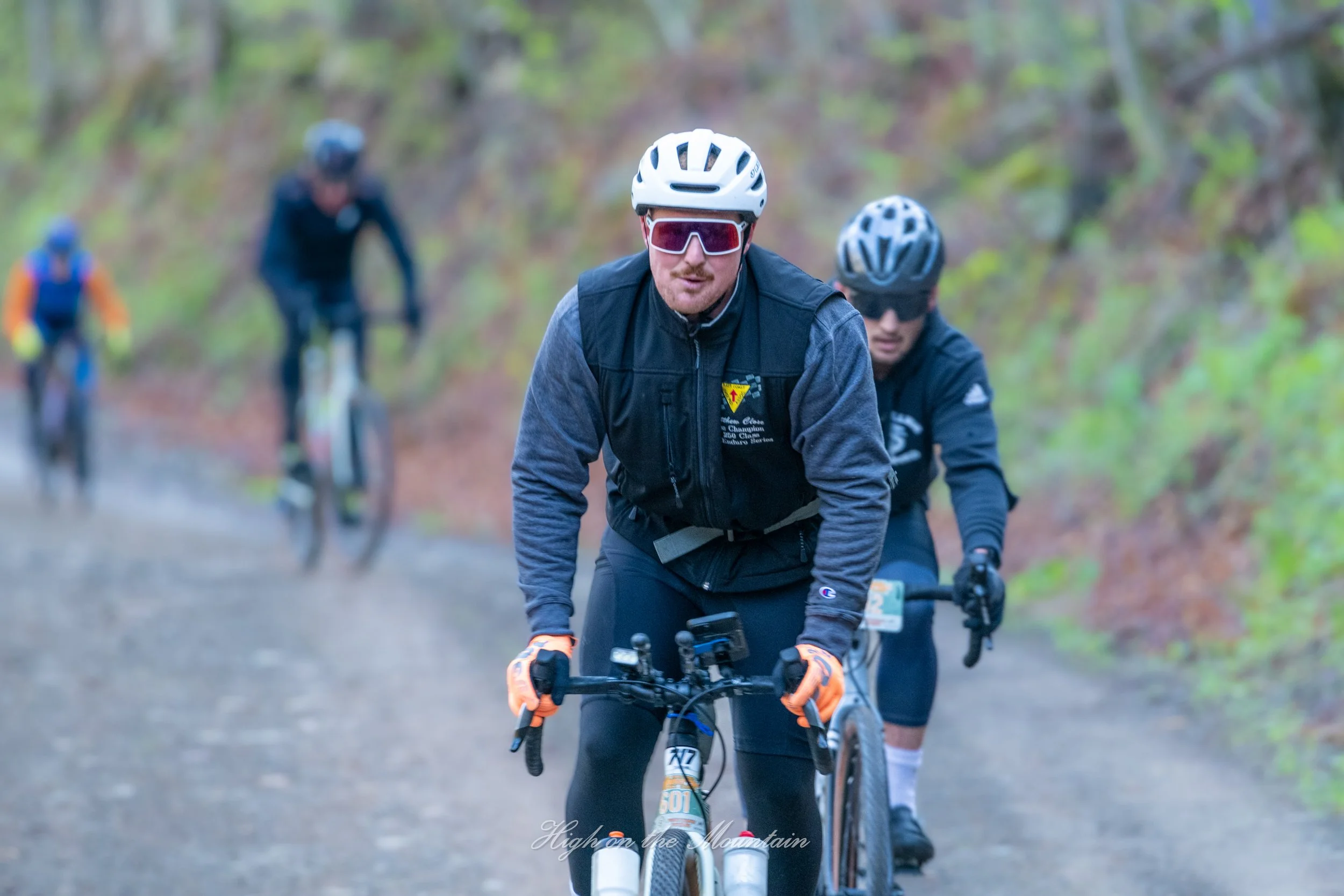 A group of people mountain biking on a trail through a forest with trees. The person in the foreground is wearing a white helmet, sunglasses, and a black vest. Other bikers are visible behind wearing helmets and outdoor gear.