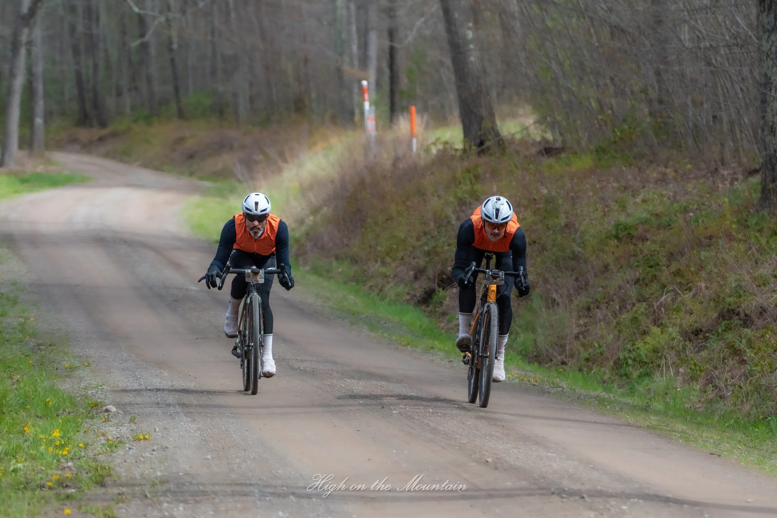 Two men wearing orange and black cycling gear and helmets riding mountain bikes on a dirt trail through a wooded area, with trees and grass on either side of the path.