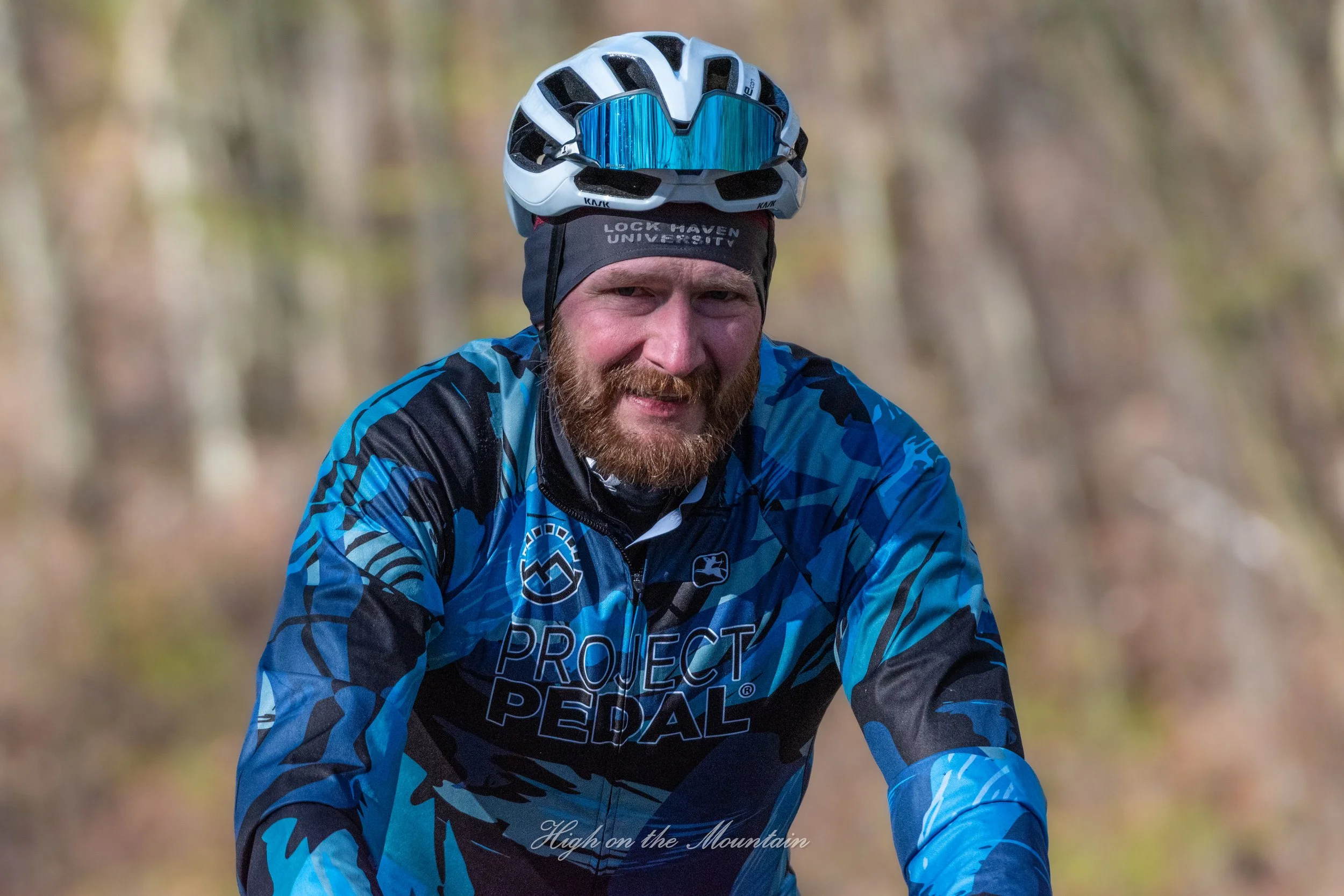 A man wearing a blue mountain biking jersey and a black headband, with a bicycle helmet and reflective sunglasses placed on his helmet, smiling outdoors with a blurred natural background.