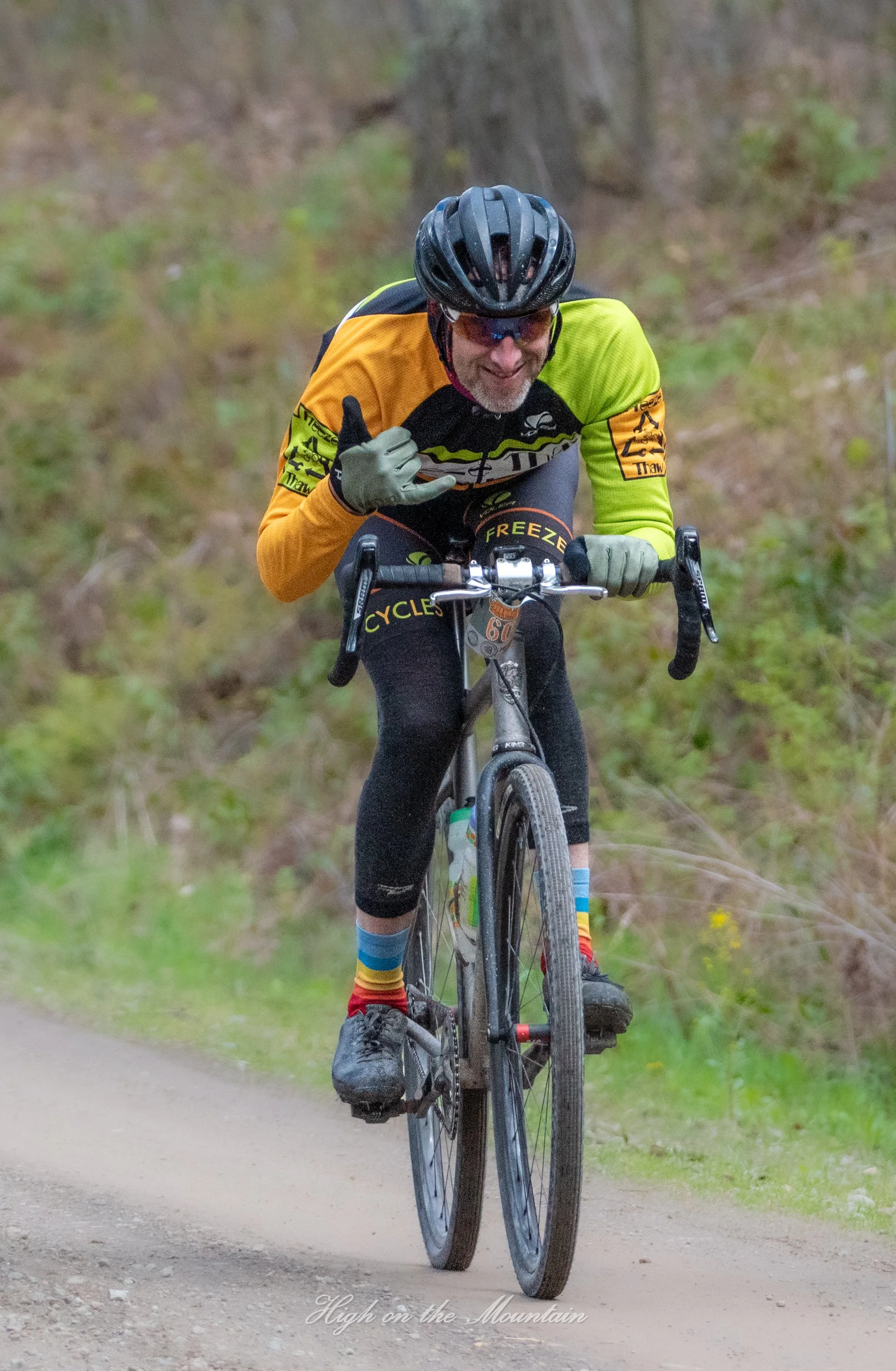 A man biking on a trail through a forest, wearing a helmet, sunglasses, and a colorful cycling jersey, giving a thumbs up and smiling.