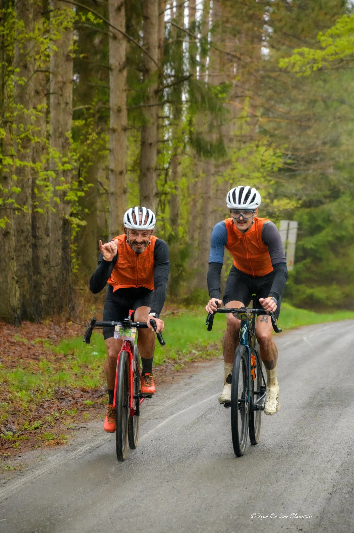 Two men riding bicycles on a road through a forested area during daytime, wearing helmets and orange vests.
