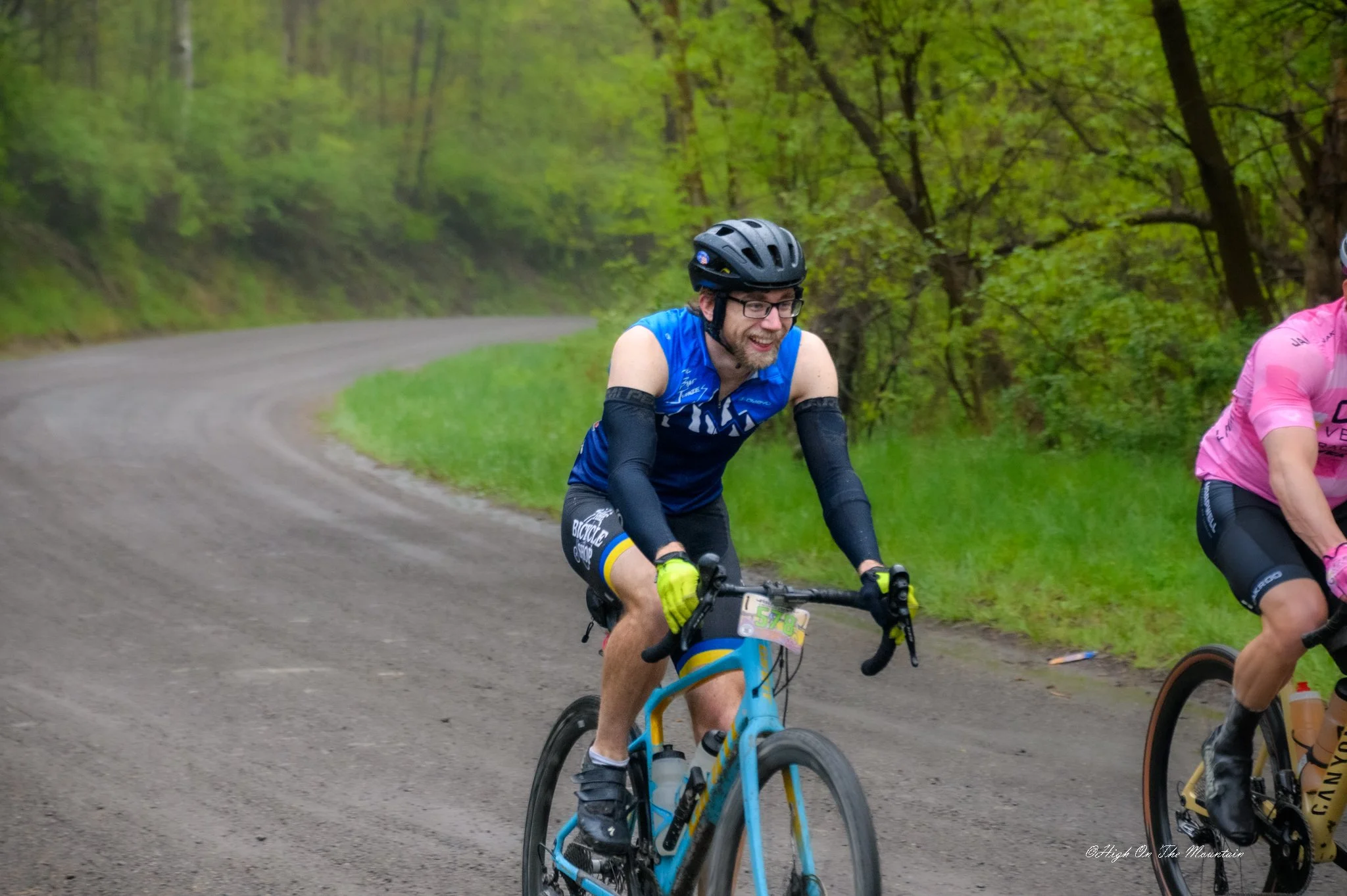 A man wearing a black helmet, glasses, and a blue sleeveless cycling jersey riding a blue mountain bike on a forest trail. He is smiling and appears to be enjoying the ride with others, partially visible, smiling beside him.