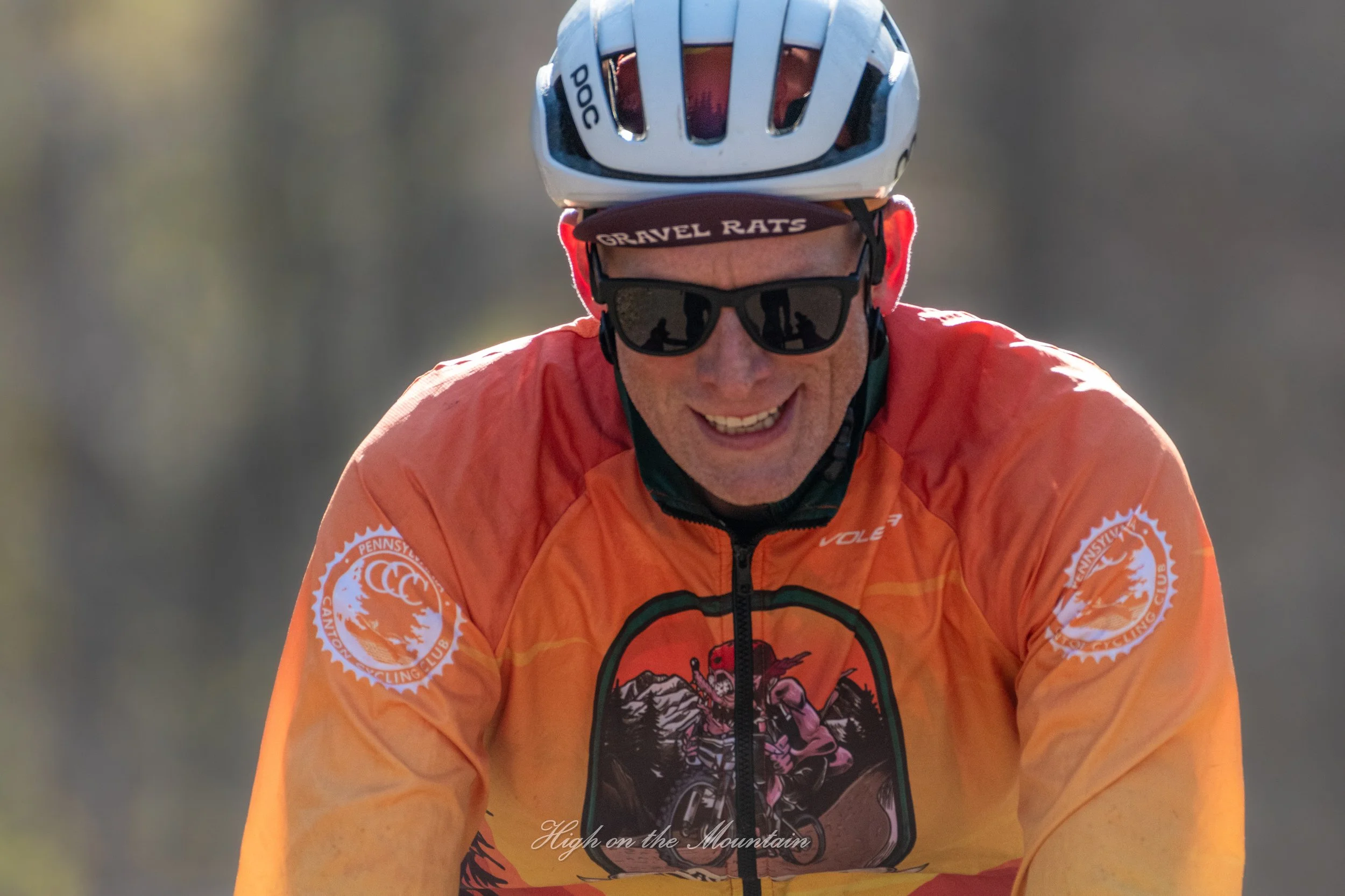 A man wearing a cycling helmet and sunglasses is smiling while riding a mountain bike. He has on an orange jacket with a logo on the sleeve, and a headband that reads 'GRAVEL RATS'. The background is blurred, indicating motion, with the text 'High on