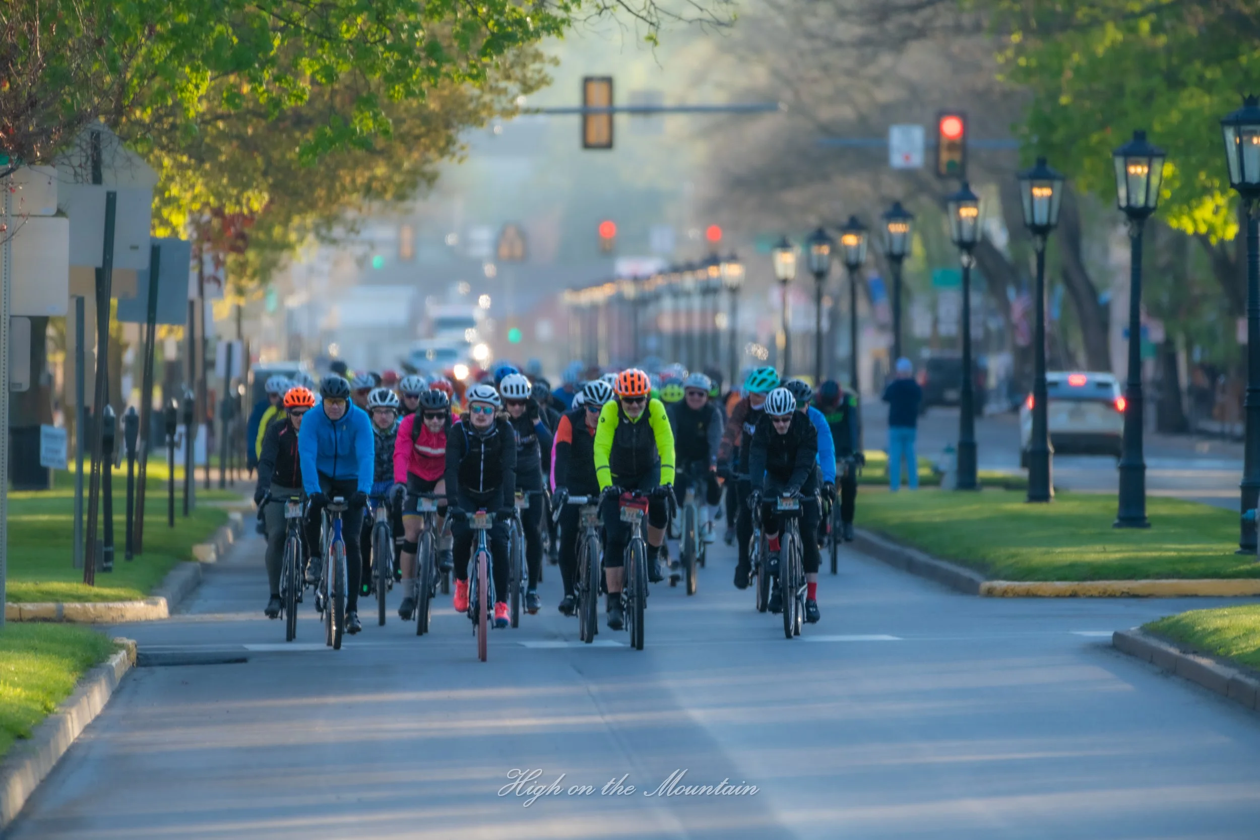 A group of cyclists riding on a city street lined with lampposts and trees, with traffic lights visible in the background.