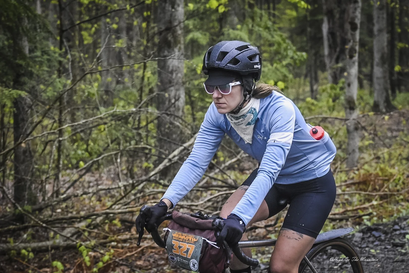 A woman cycling on a muddy trail in a forest, wearing a black helmet, sunglasses, a blue long-sleeve cycling jersey, black shorts, and gloves, with mud splattered on her legs.