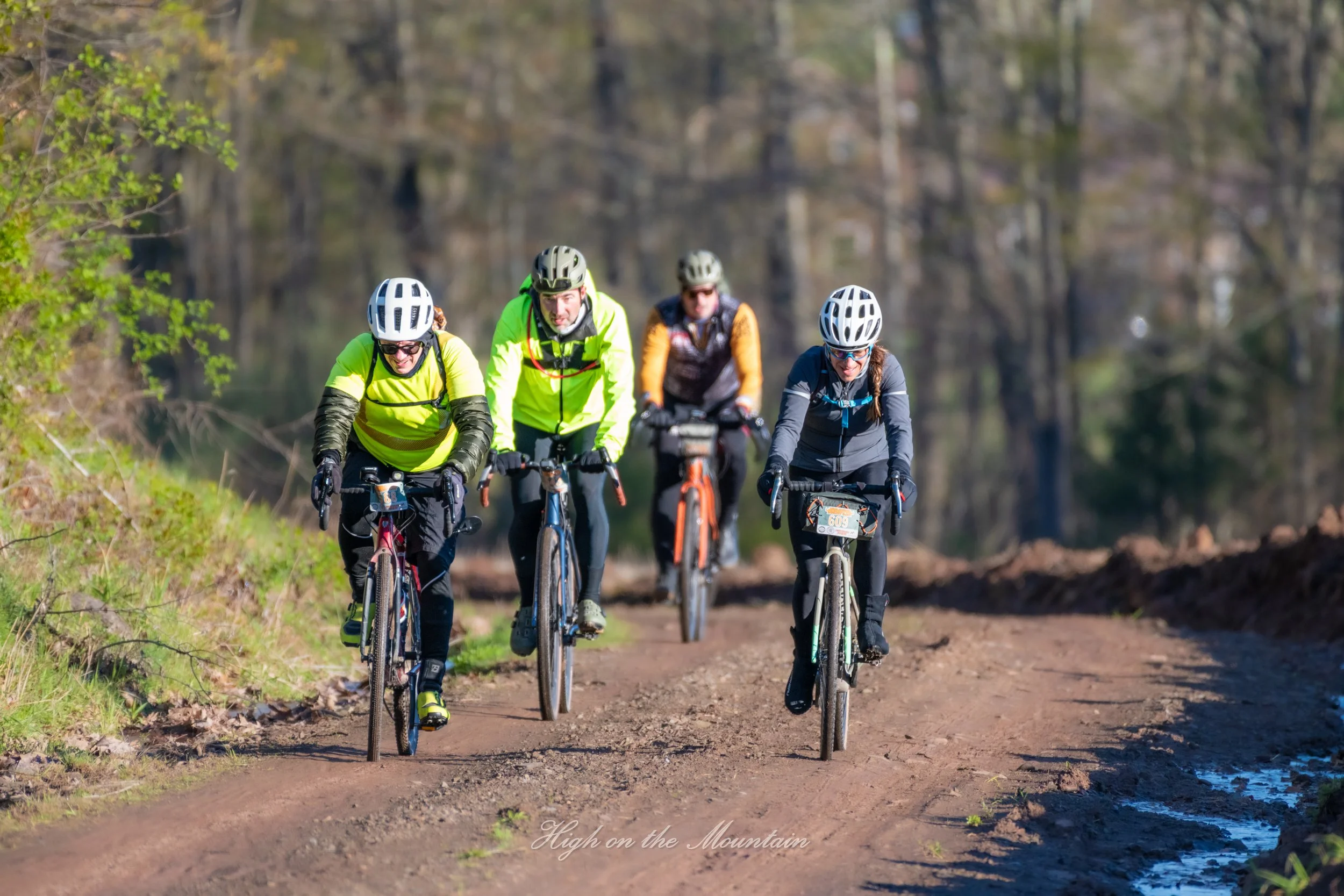 Four people riding mountain bikes on a dirt trail in a wooded area, wearing helmets and bright clothing, with trees and sunlight in the background.
