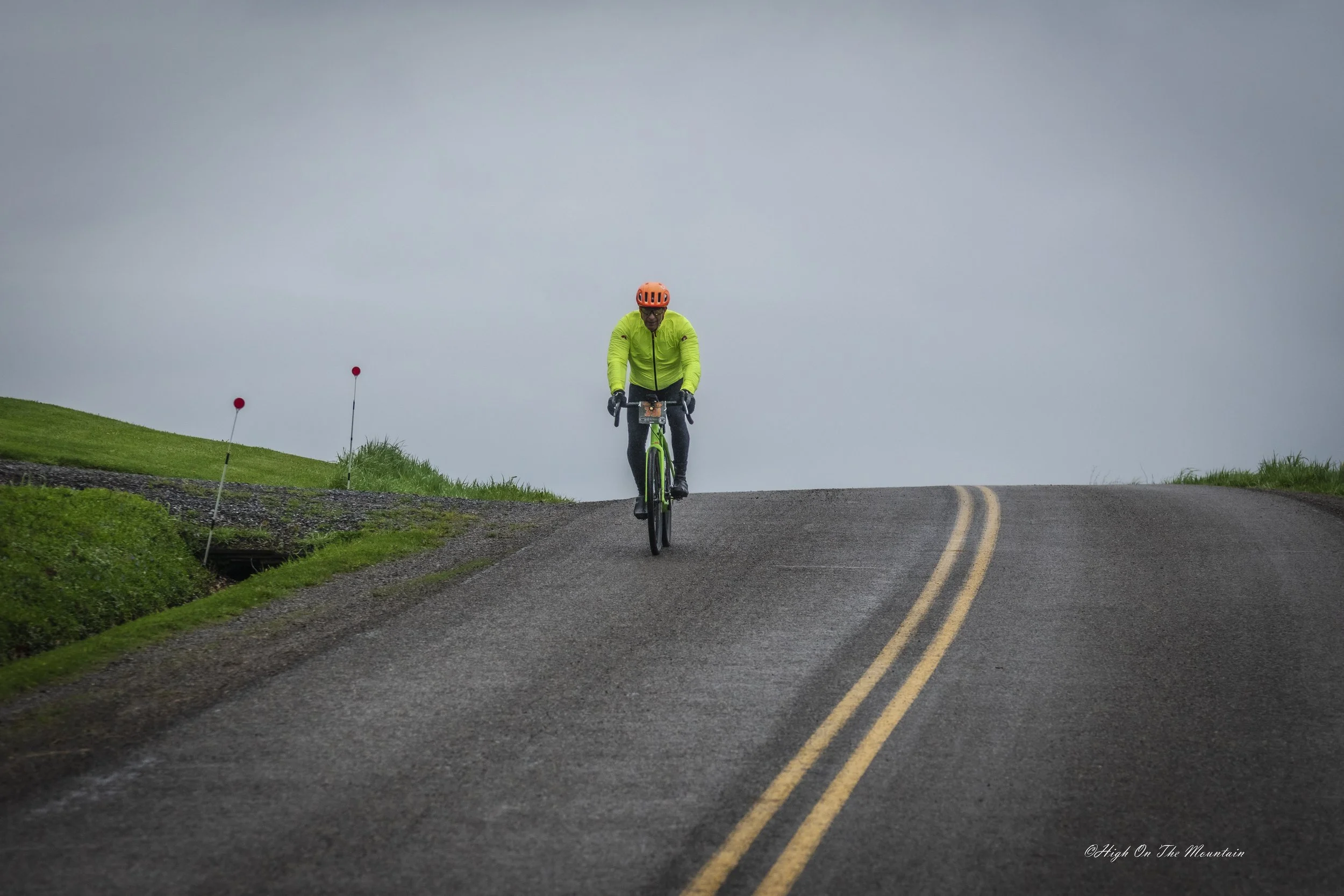 A person wearing a bright yellow jacket and orange helmet riding a bicycle on a winding mountain road with grassy sides and red markers, under a cloudy sky.