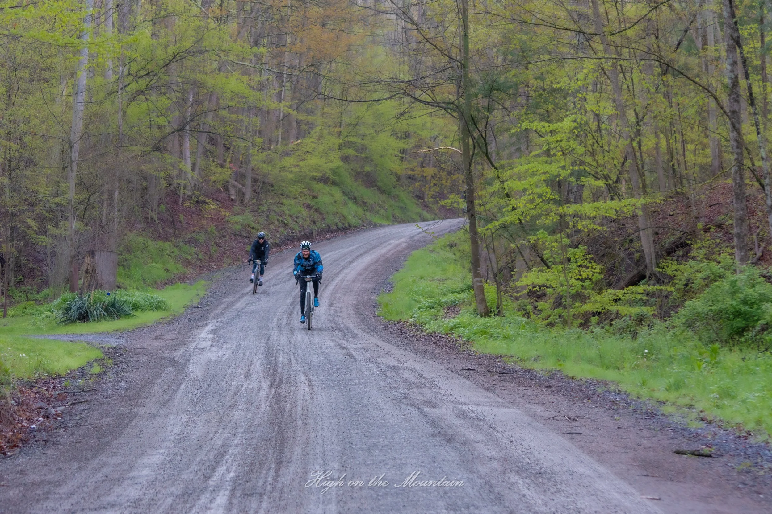 Two people riding bicycles on a gravel mountain trail surrounded by trees with new spring green leaves, overcast sky, with the words "High on the Mountain" at the bottom.