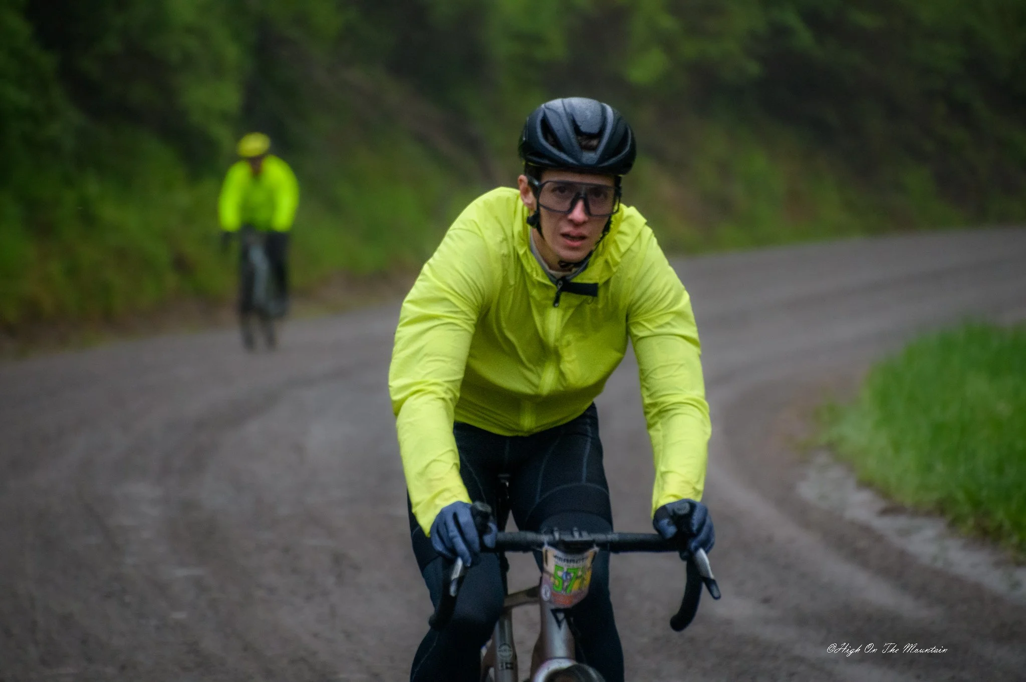 A man in a yellow jacket and black helmet riding a mountain bike on a trail surrounded by greenery, with a person in a neon yellow jacket and hat in the background.