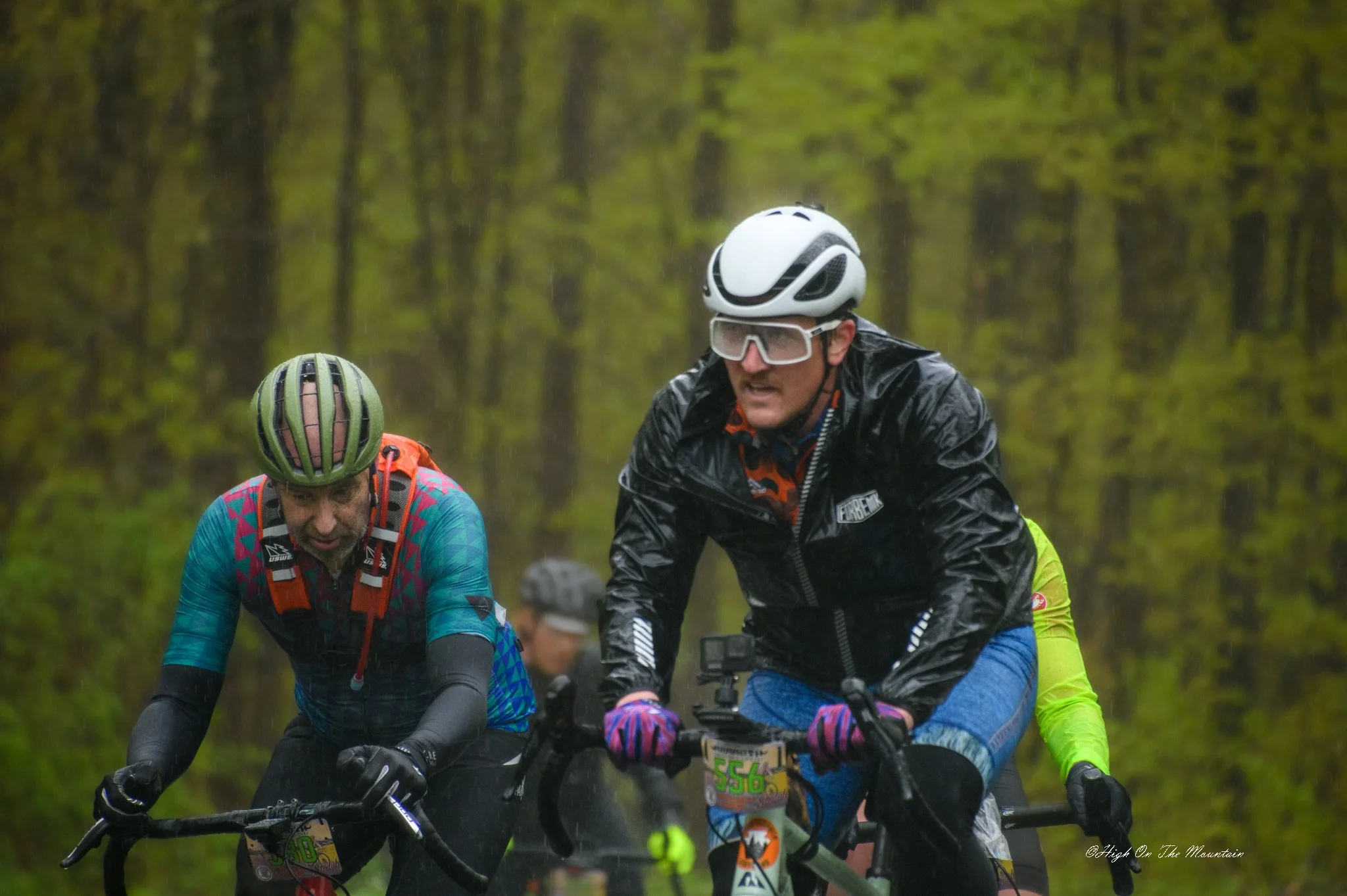 Group of mountain bikers riding on a trail through a dense green forest in the rain, wearing helmets, rain gear, and goggles.