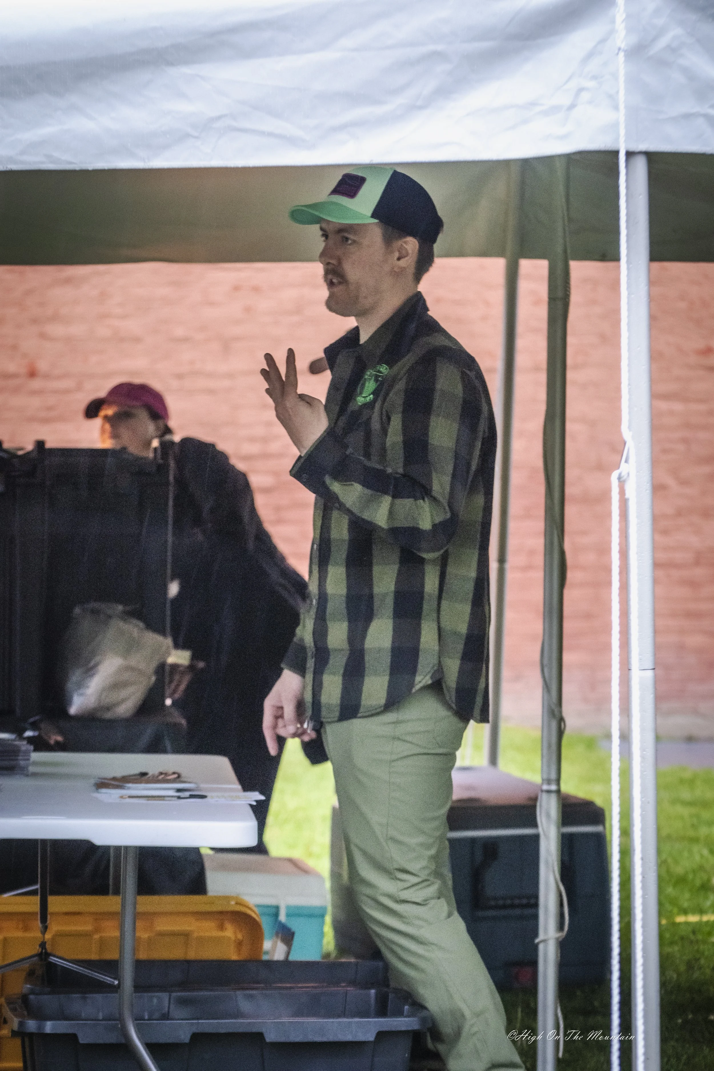 A man wearing a green and black trucker hat, a plaid shirt, and green pants standing under a tent, engaged in conversation during an outdoor event.
