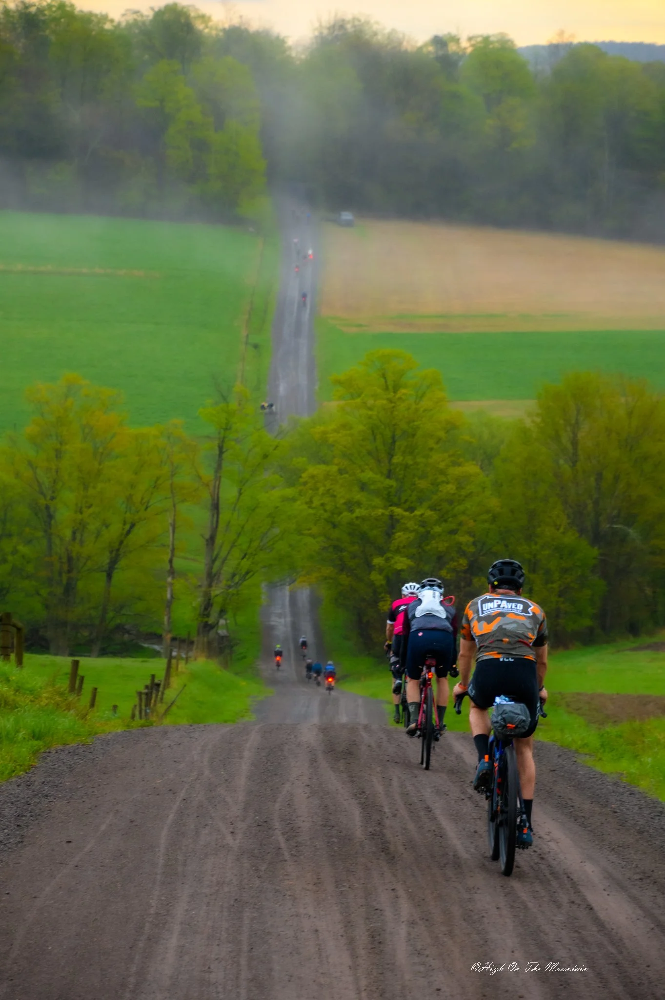 Bicyclists riding on a dirt road through countryside with green fields and trees, with a line of cars visible on the road ahead and mist in the background.