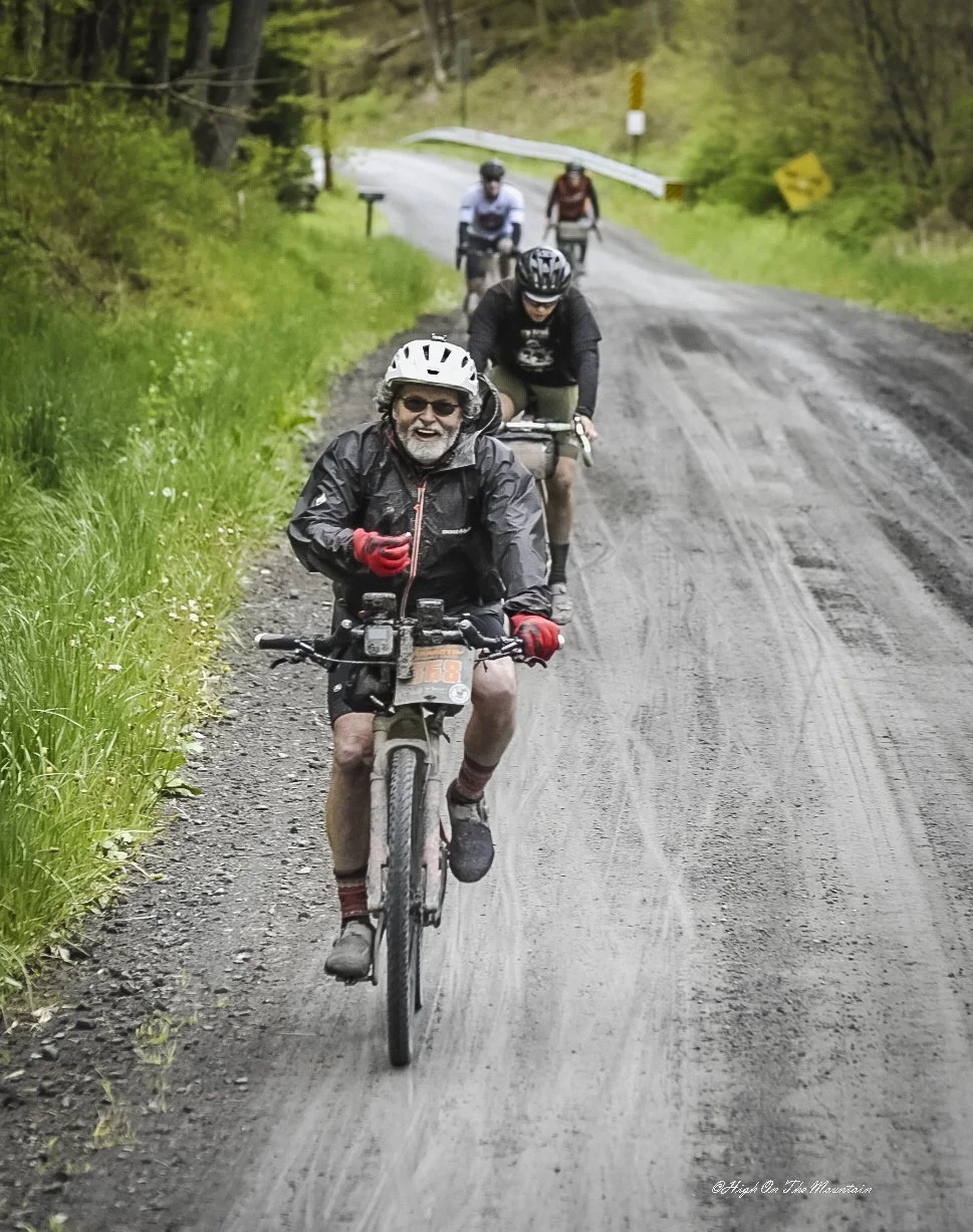A group of four people biking on a dirt trail through a green, wooded area, with the person at the front wearing a helmet, glasses, and a black jacket, smiling.