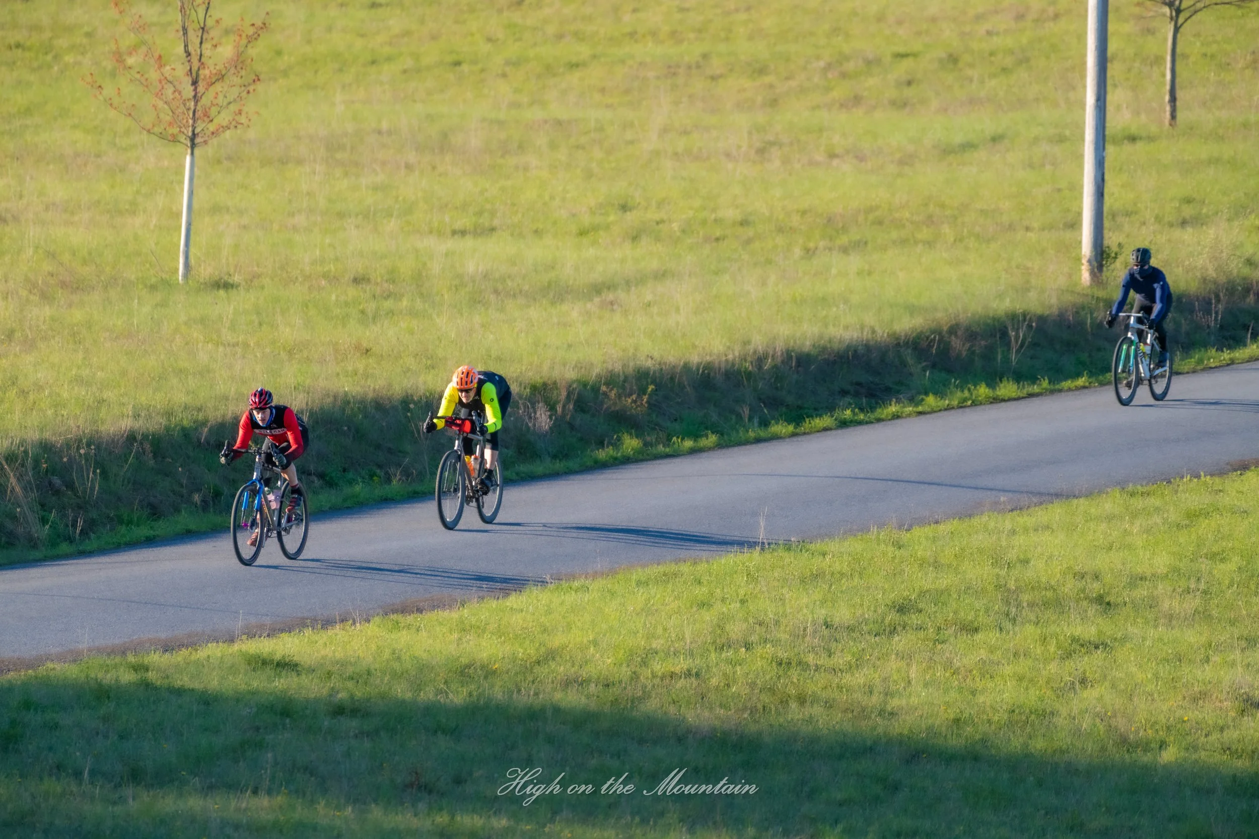 Three mountain bikers riding on a paved trail through a grassy hillside with trees, under a clear sky.