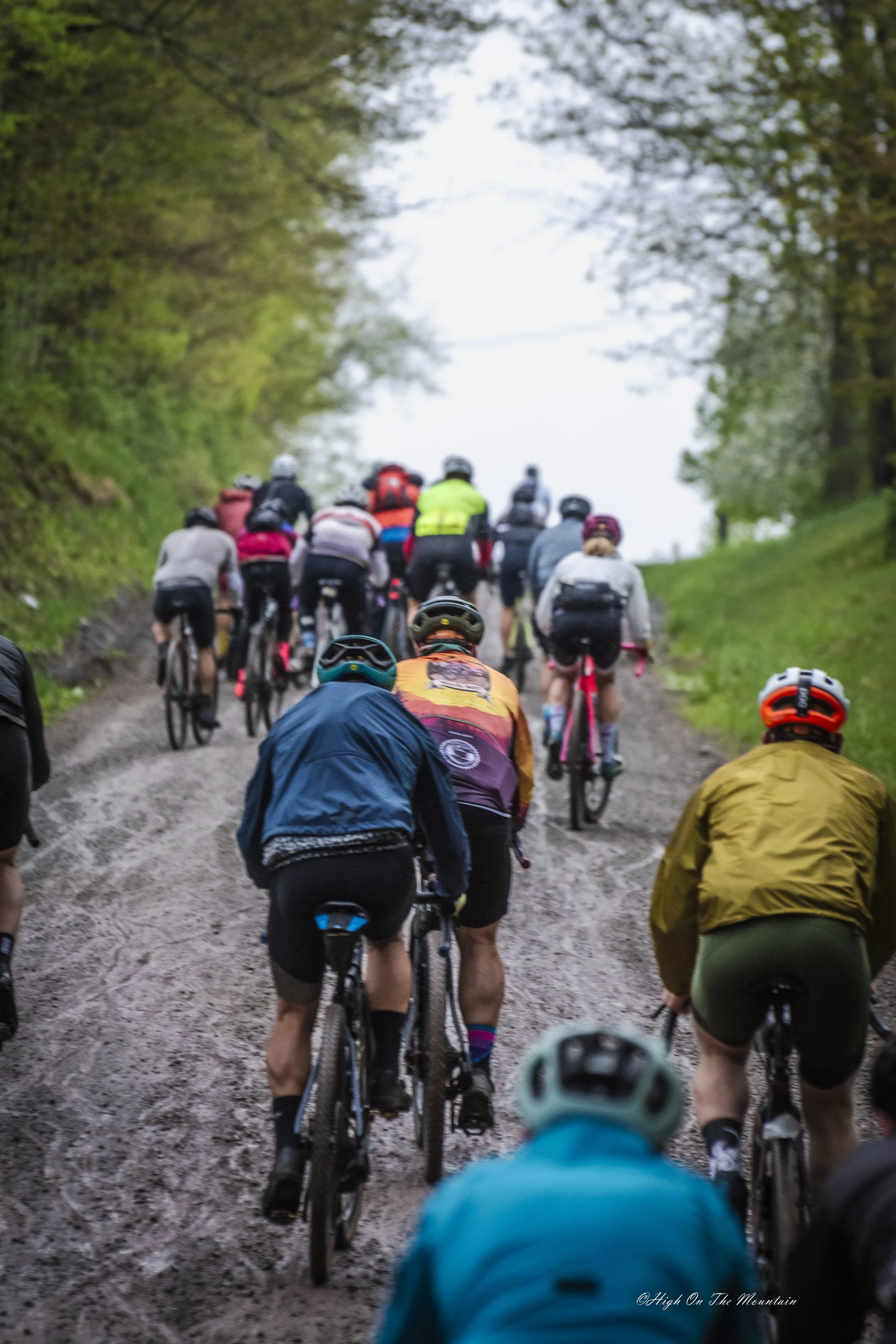 Group of cyclists riding on a muddy, tree-lined trail in daytime. Some wear helmets and colorful clothing.