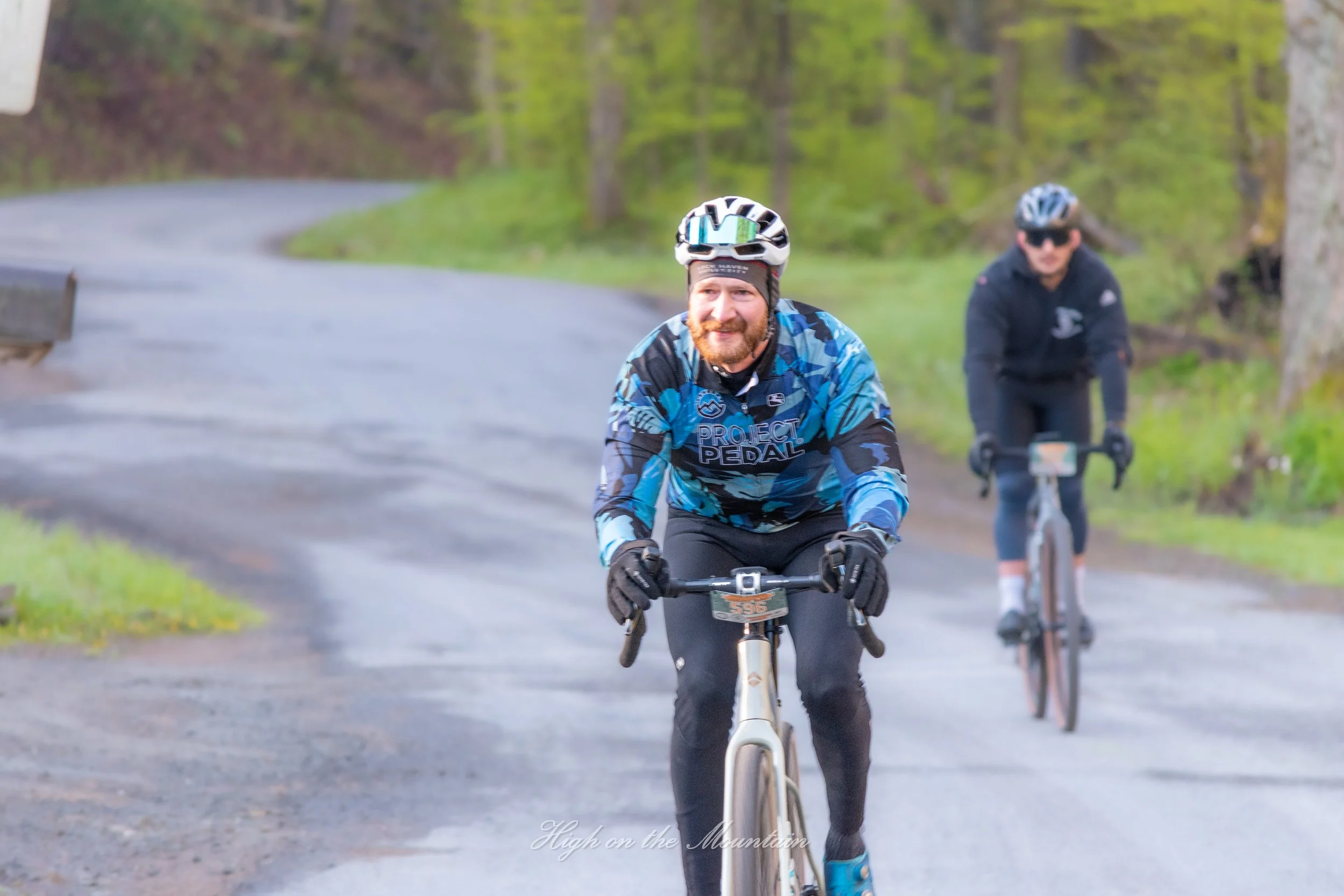 Two men riding bicycles on a paved road through a forested area with green trees. The man in front is smiling and wearing a blue patterned cycling jersey, helmet, and sunglasses. The man behind is wearing a black helmet, sunglasses, and a dark jacket