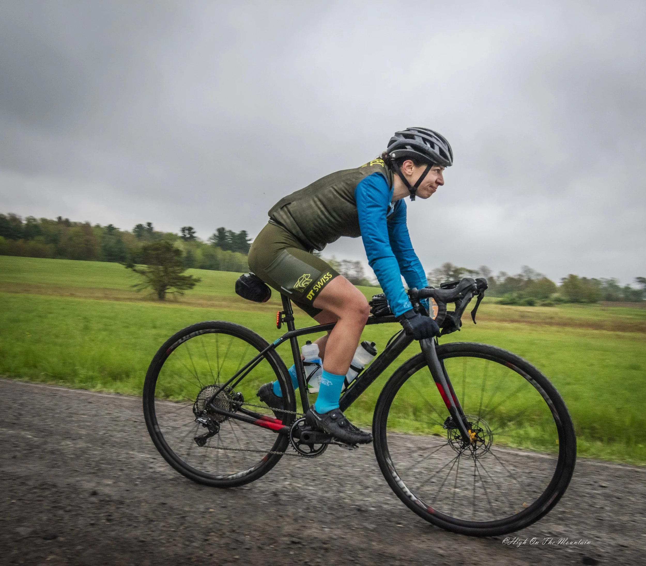 A person riding a mountain bike on a paved path through a grassy area with trees in the background on a cloudy day. The cyclist is wearing a black helmet, a green vest, and blue sleeves.