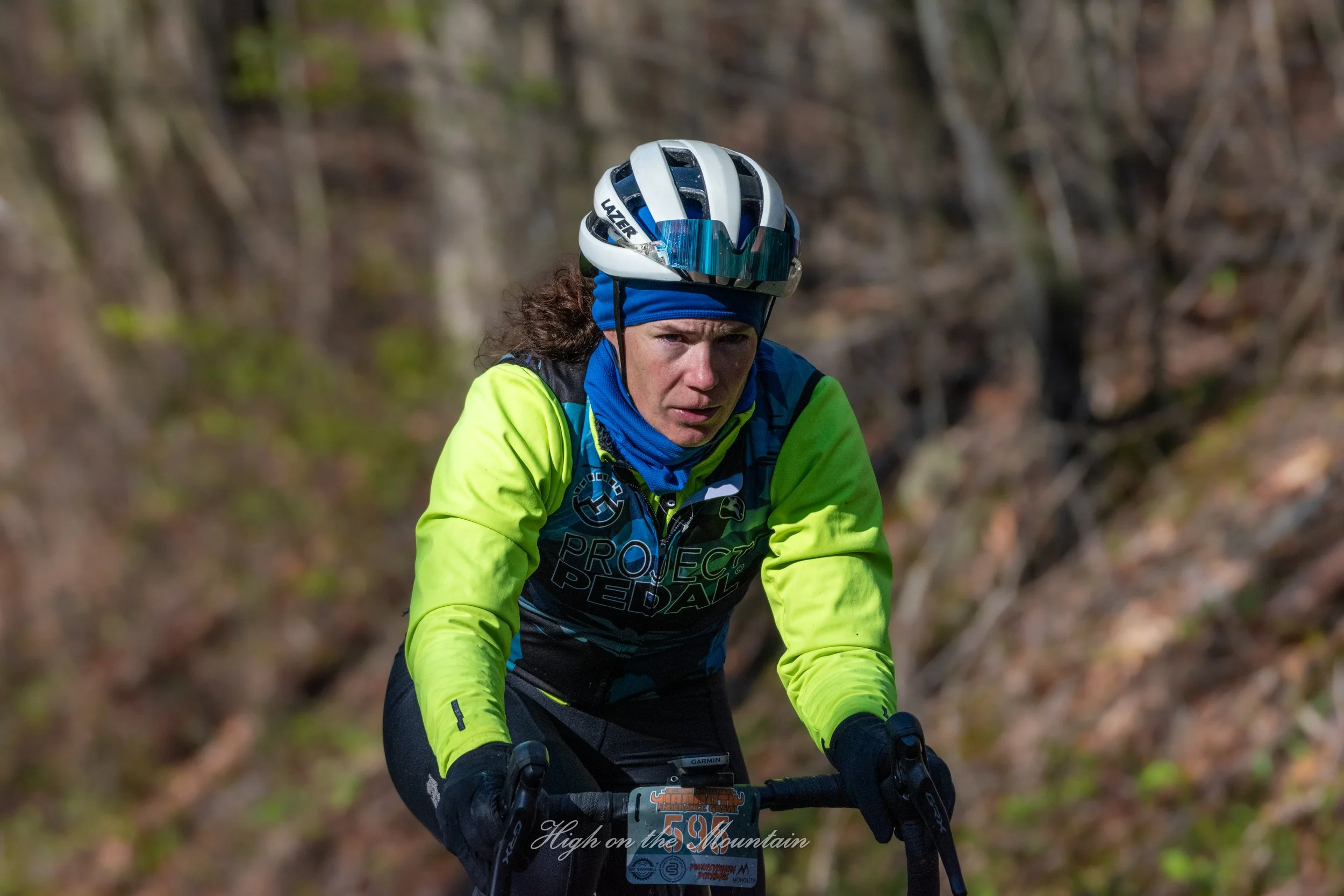 A cyclist wearing a helmet, blue headband, and neon yellow jacket riding mountain bike on trail surrounded by trees during daytime.