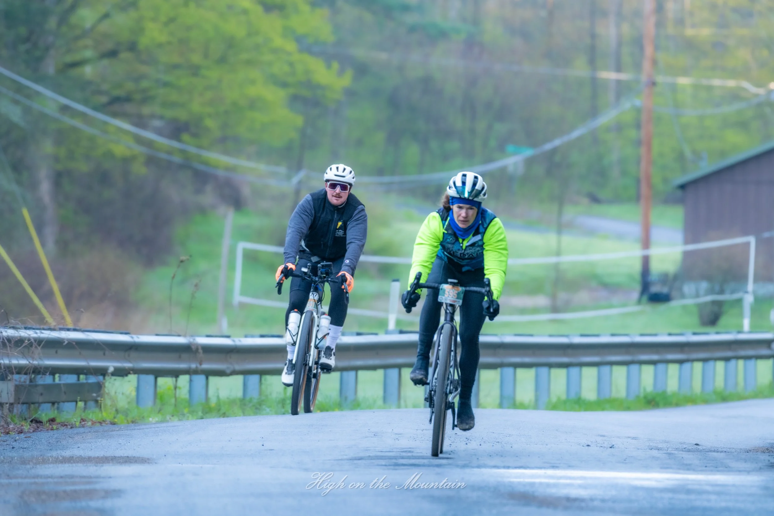 Two cyclists riding mountain bikes on a paved path surrounded by lush green trees. The cyclists are wearing helmets and athletic clothing, with one in a black and gray outfit and the other in a bright yellow and blue jacket. They appear focused and d