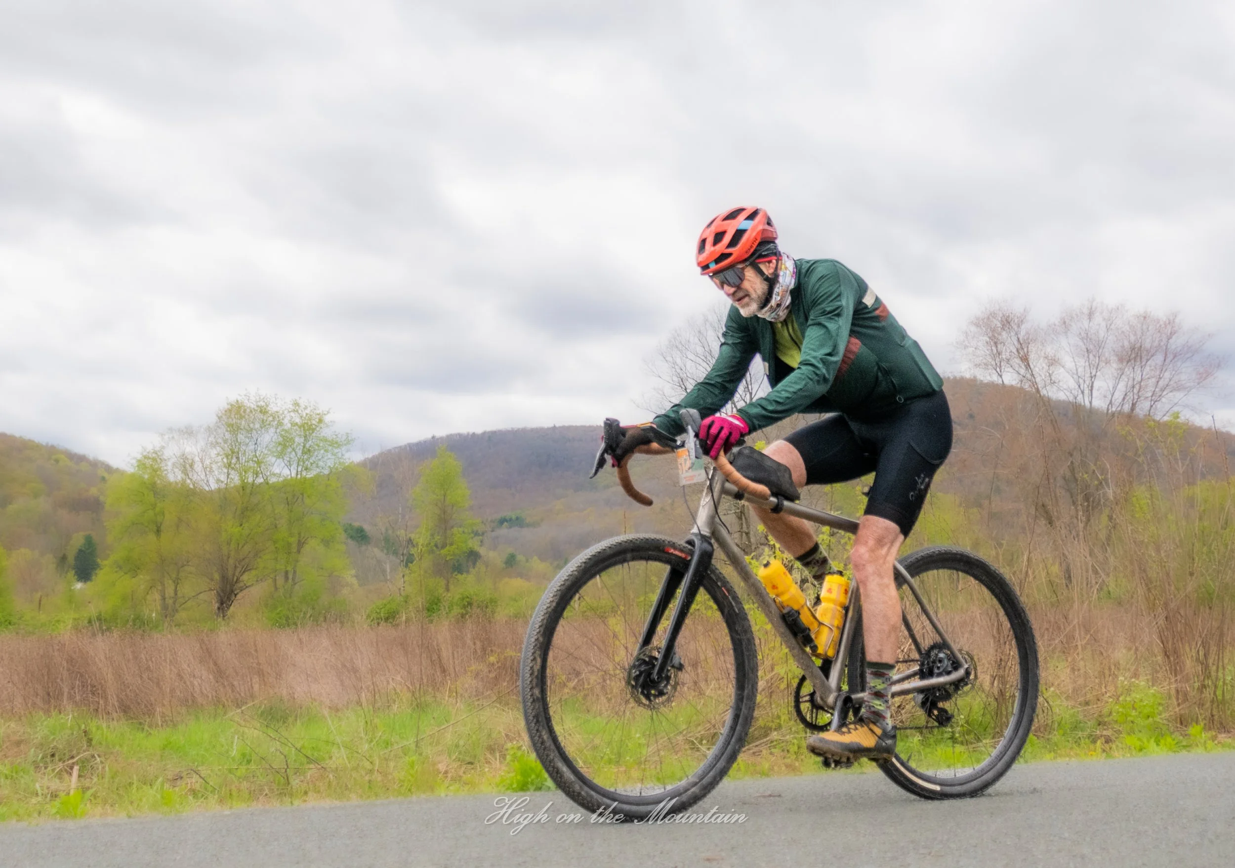 A man wearing a red helmet, sunglasses, a green jacket, black shorts, and pink gloves is riding a mountain bike outdoors on a cloudy day. The landscape features grassy fields and distant hills with trees.