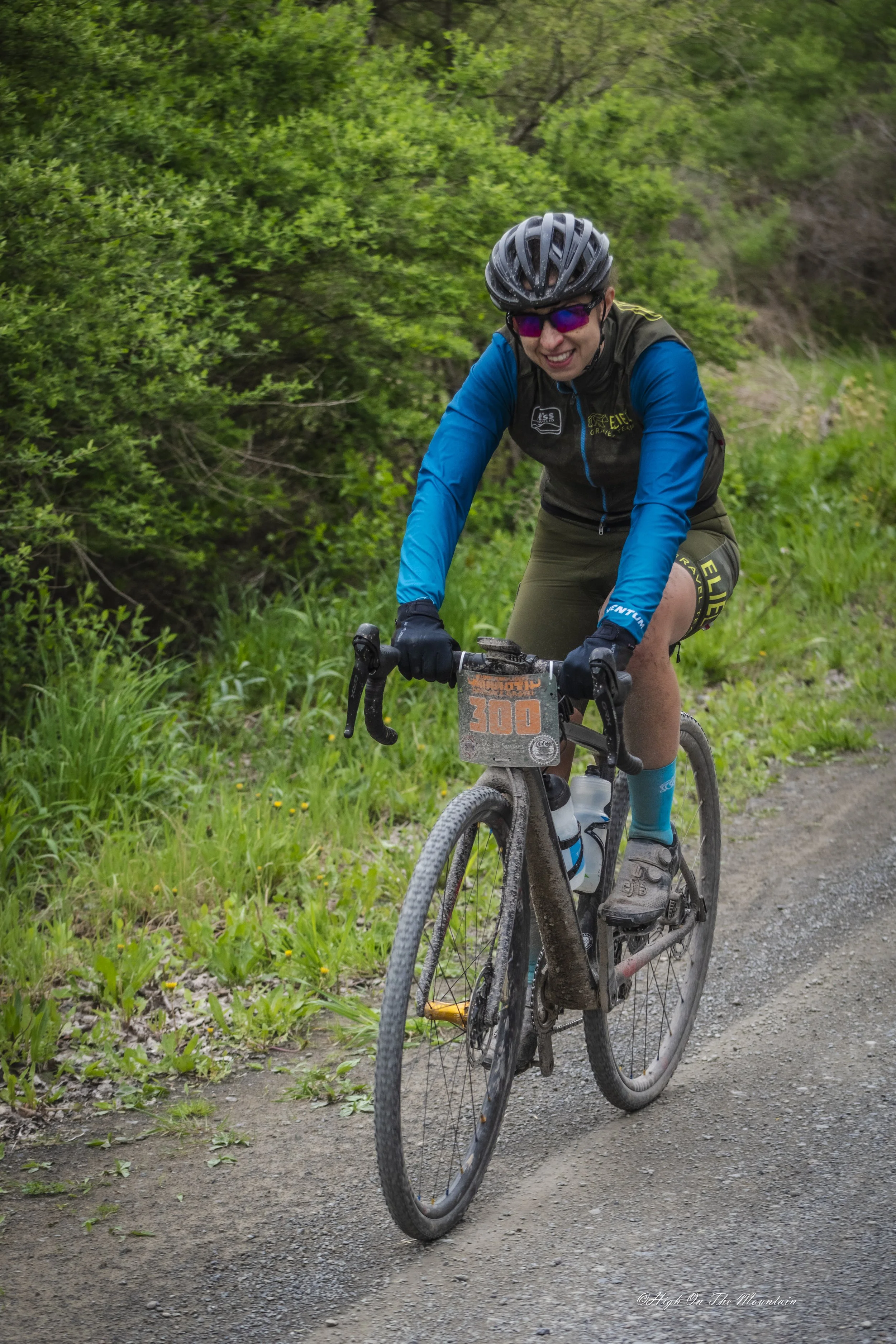 A person riding a mountain bike on a gravel trail surrounded by green foliage. The rider is wearing a black helmet, sunglasses, blue and black cycling gear, and smiling.