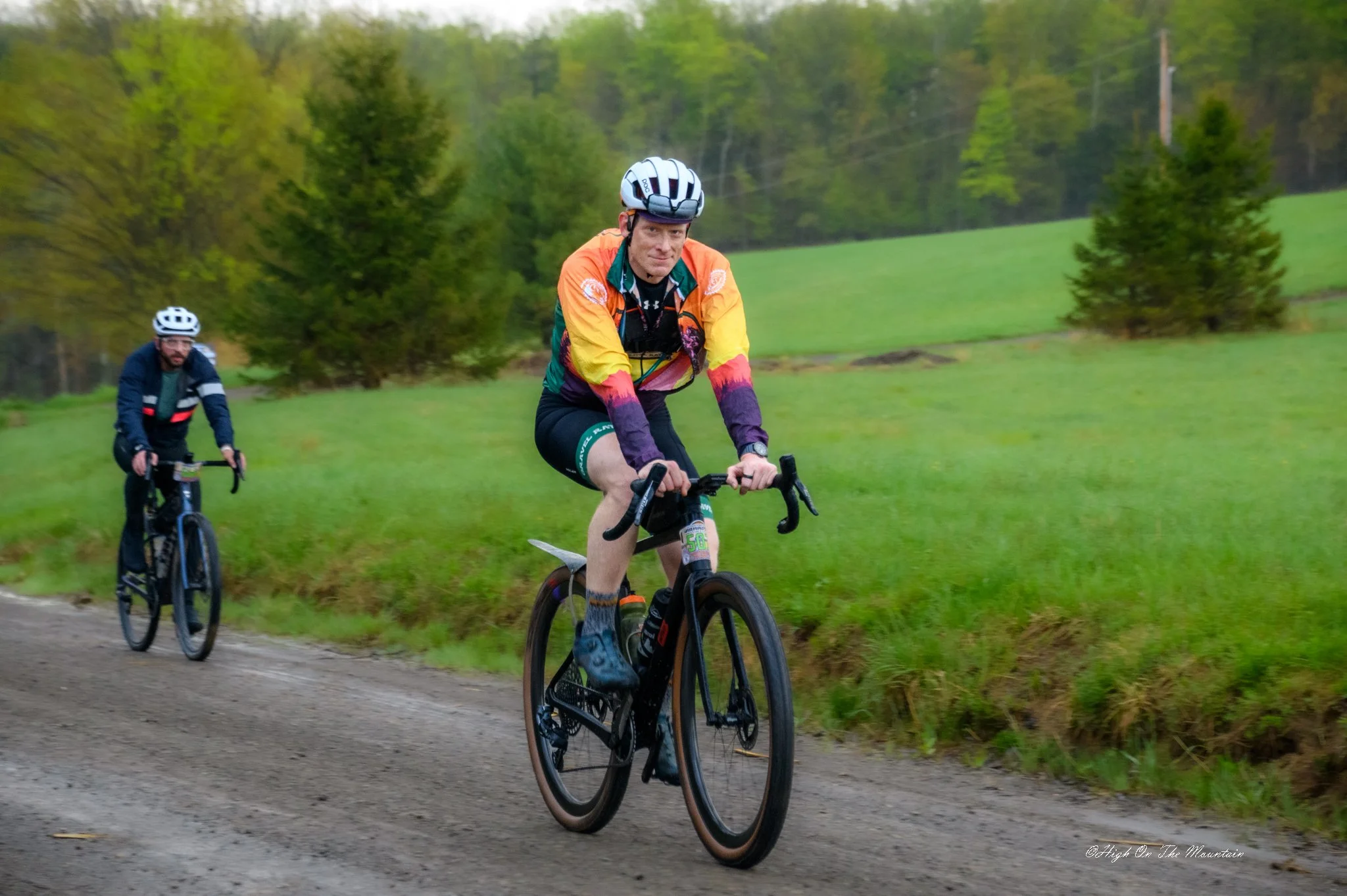 Two men riding mountain bikes on a dirt trail through a lush green landscape with trees and rolling hills in the background.