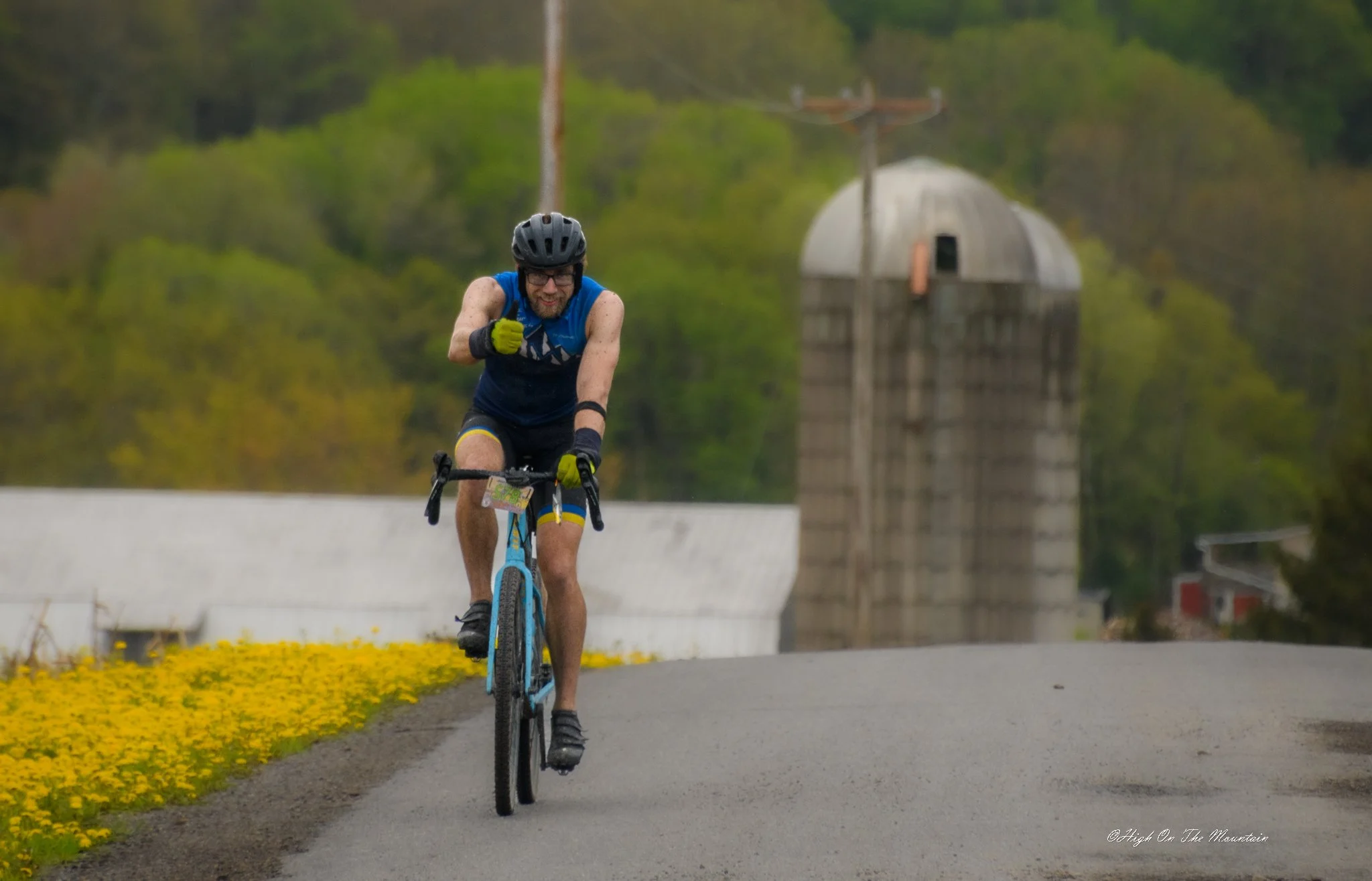 A man biking on a paved path in a rural area with trees and a small wooden building in the background. He is wearing a helmet, glasses, and a blue athletic outfit, and is smiling while riding.