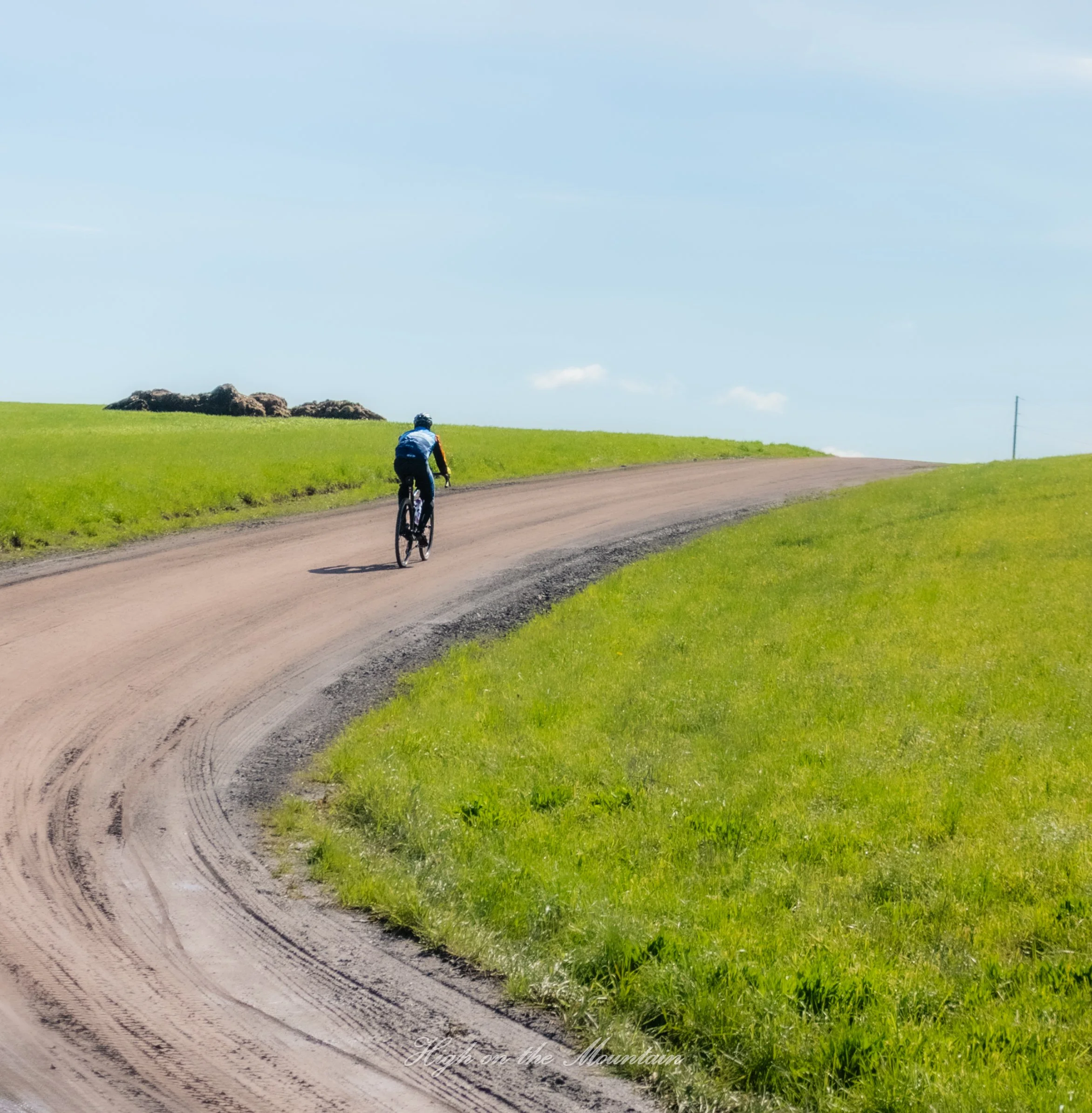 A person riding a bicycle on a dirt path through green grass under a blue sky with some clouds.