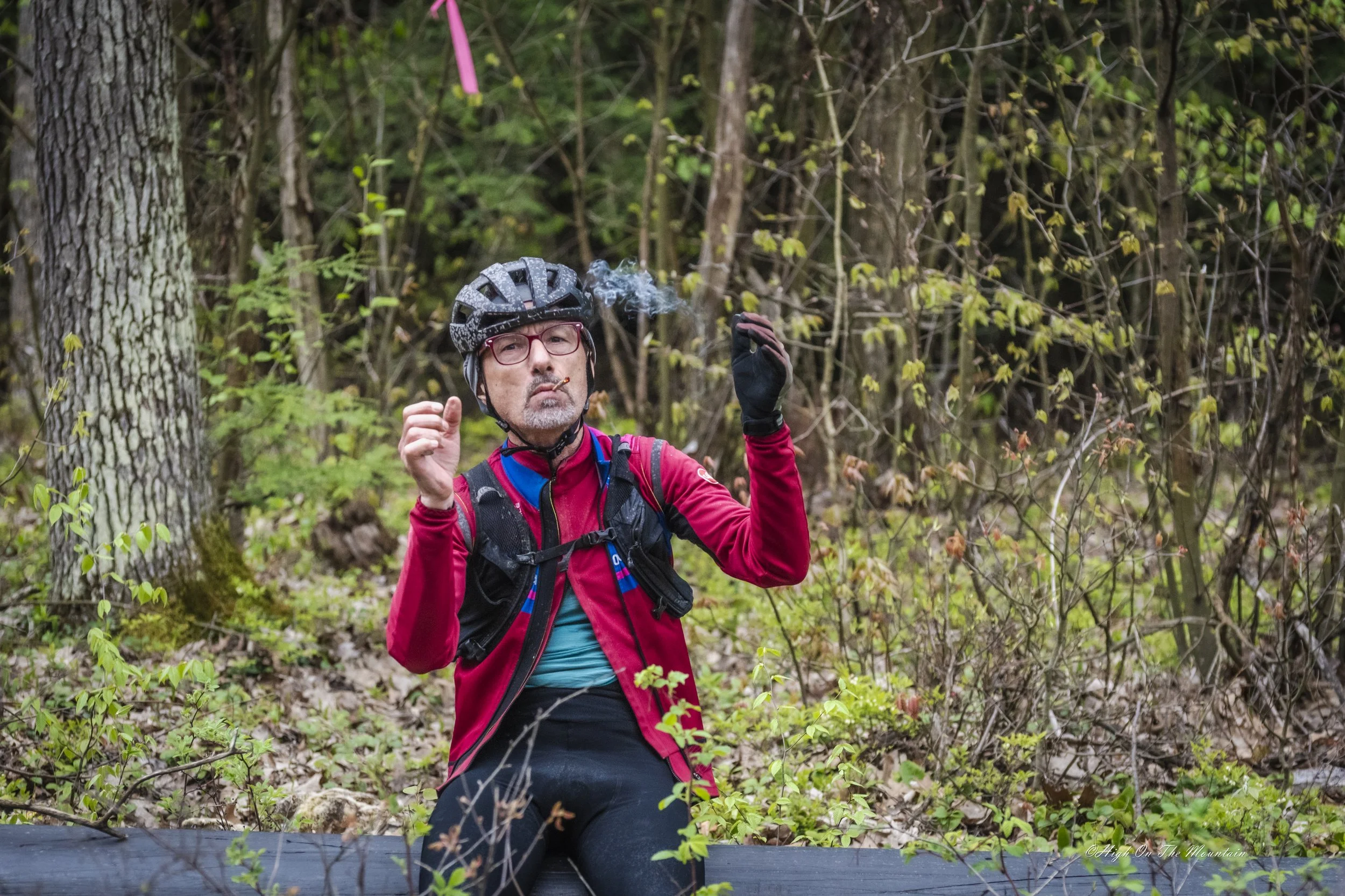 A middle-aged man with gray hair and glasses, wearing a black bicycle helmet, red jacket, and gloves, sitting on a fallen tree in a dense forest, smoking a cigarette.