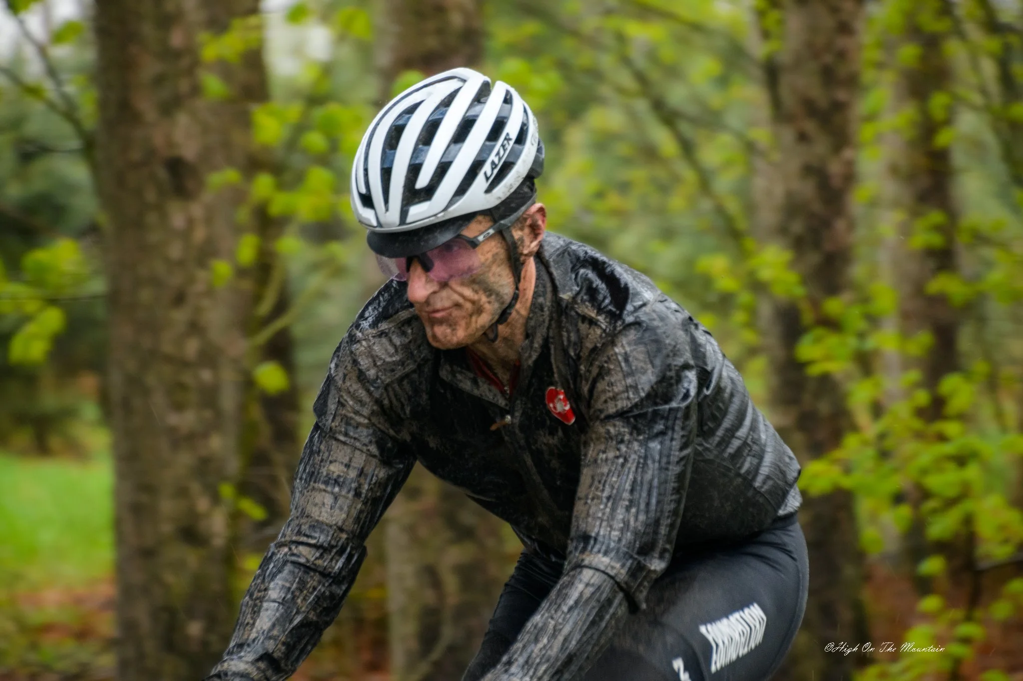 A man cycling through a green forest, wearing a black patterned jacket, black pants, a white helmet, and pink sunglasses, covered in mud.